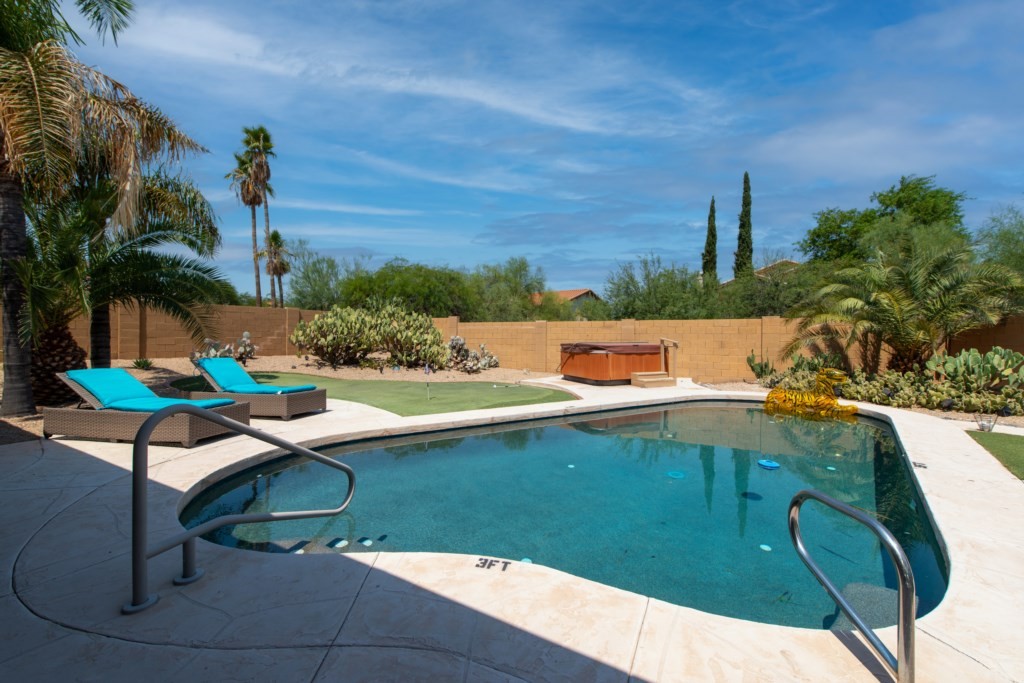 A serene backyard features a crescent-shaped swimming pool surrounded by a stone patio with lounge chairs under a clear blue sky, offering an inviting space for relaxation at an affordable luxury vacation rental house.