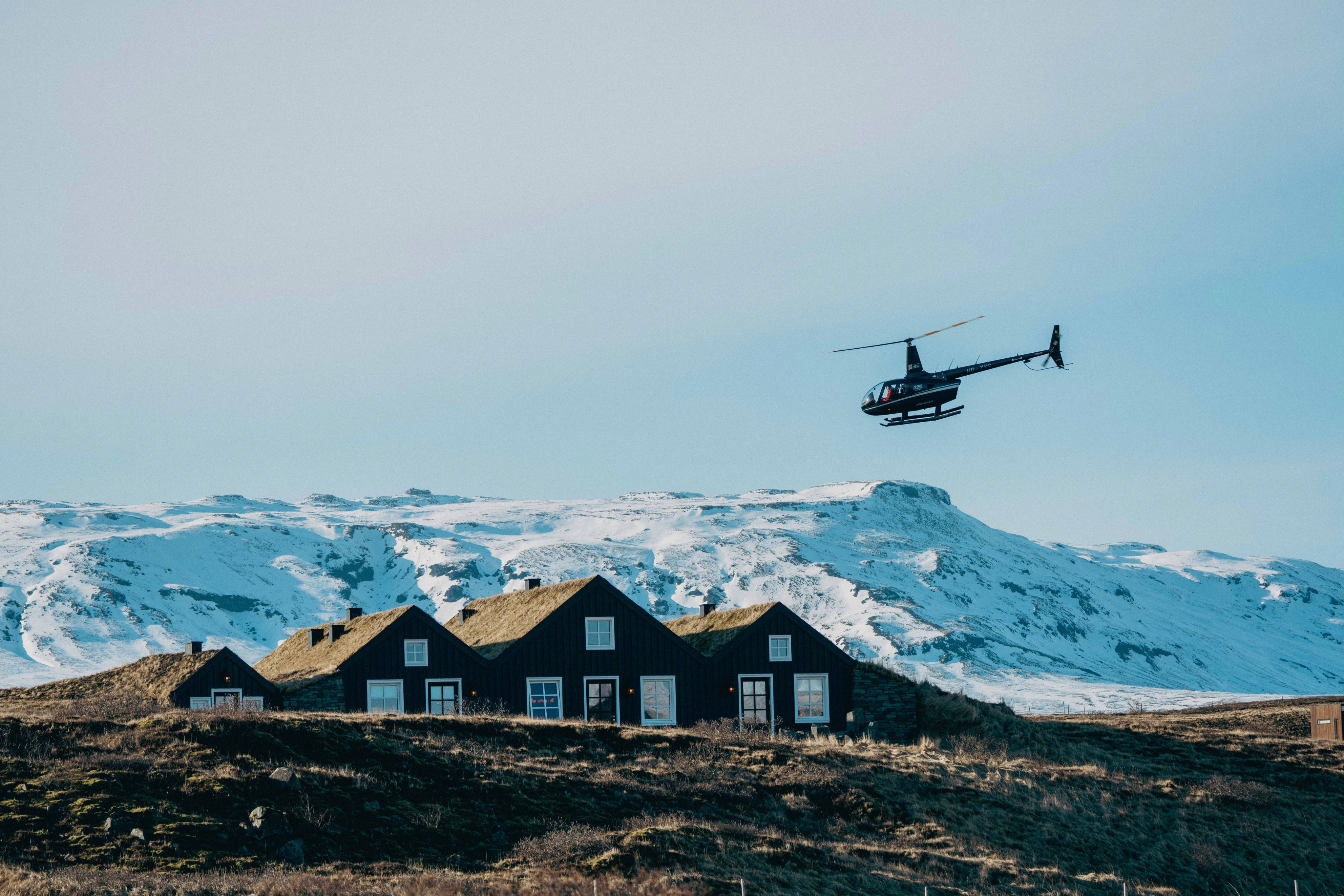 A helicopter flies over black turf-roofed cabins with snow-capped mountains behind