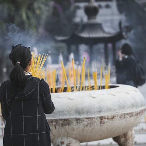 Una persona accende bastoncini d'incenso in un grande bruciatore d'incenso all'aperto in un tempio, con un'altra persona sullo sfondo.