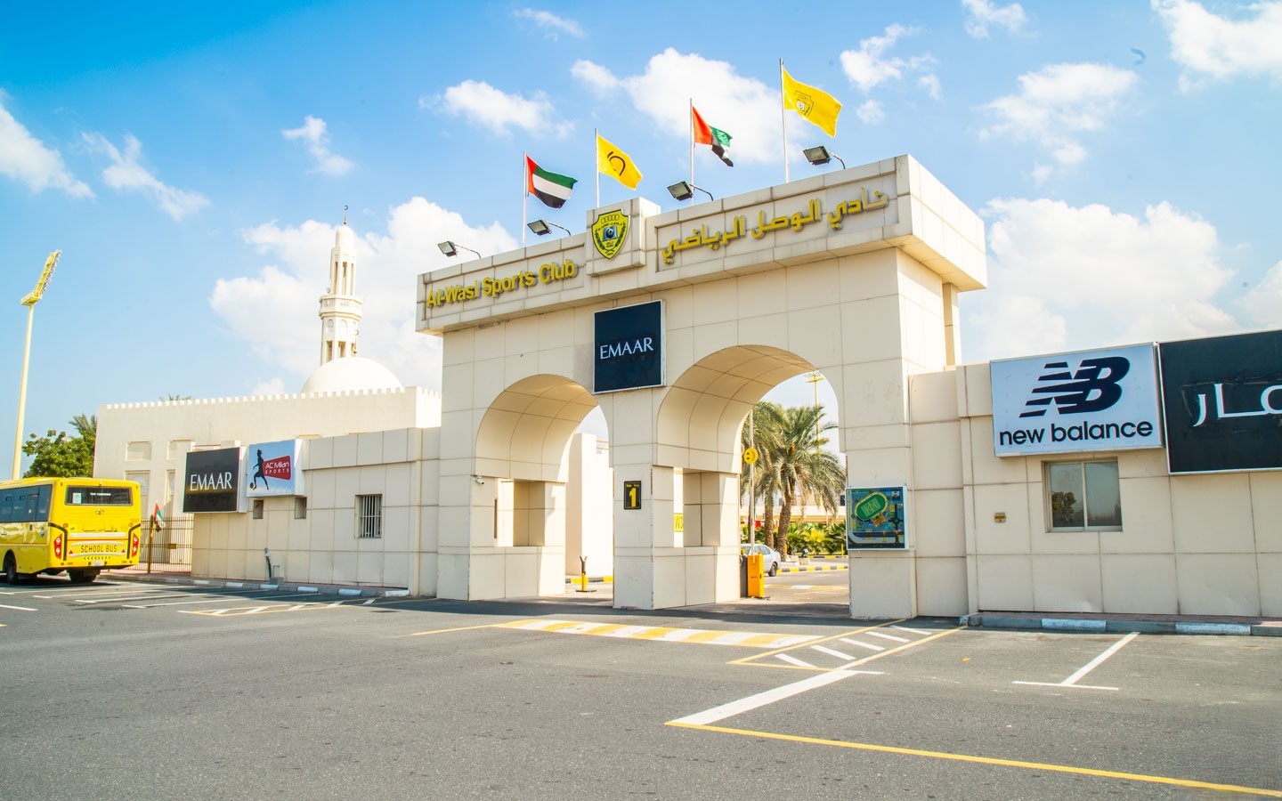 The arched entrance gate of the Al-Hewas Sports Club, topped with flags and Emaar/New Balance branding, near a mosque minaret.