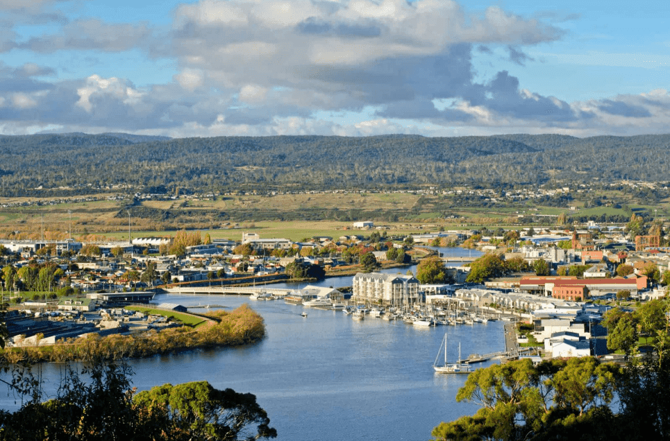 Coastal town with a river and marina, mountains in background.