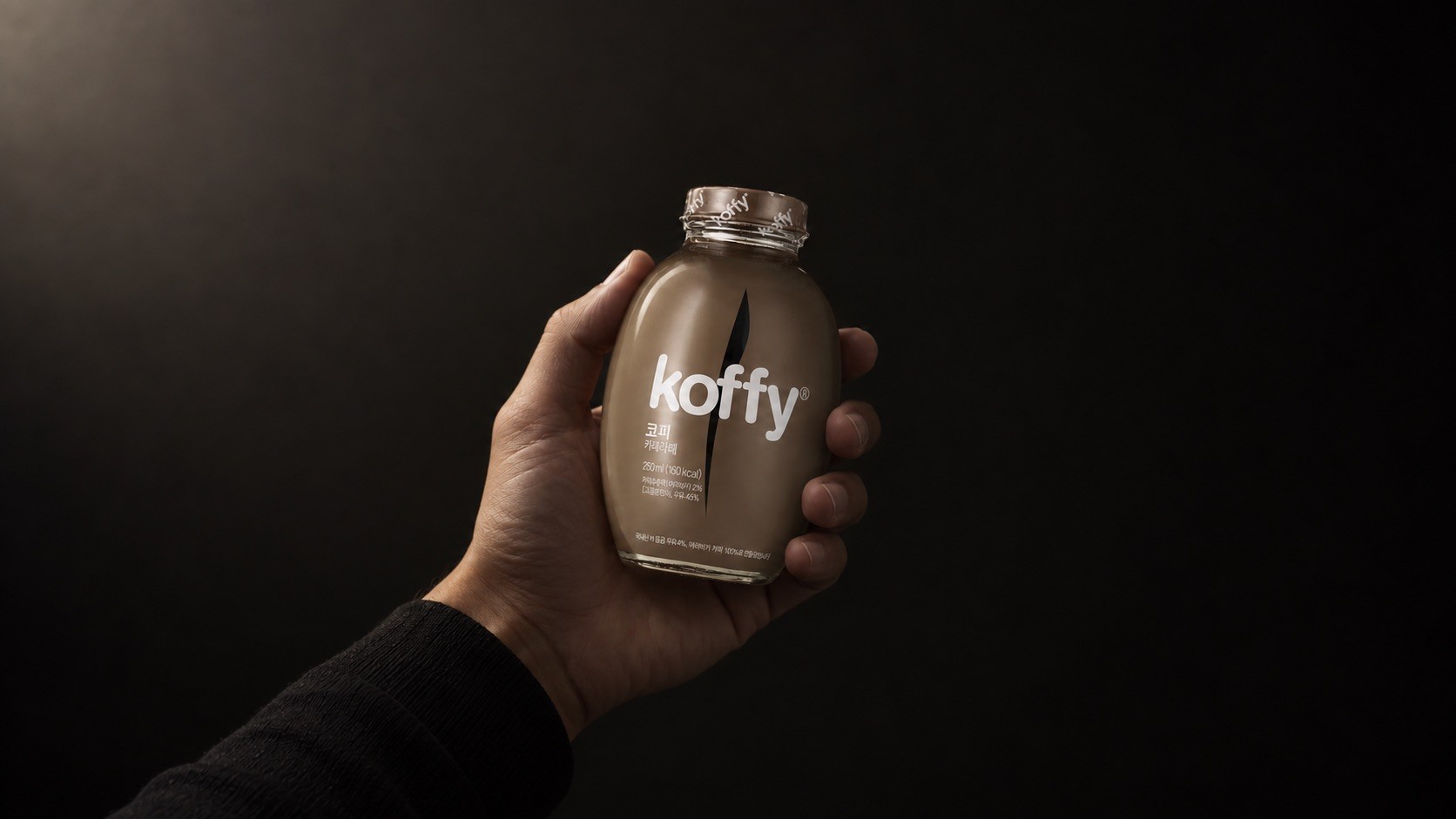 A hand holding a Koffy bottle against a dark background, lit from above.