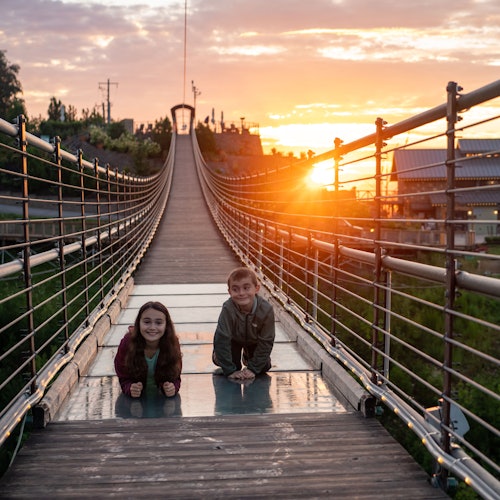שני ילדים שוכבים על גשר תלוי בזמן שקיעה, מחייכים למצלמה כשברקע יש בניינים ועצים.
