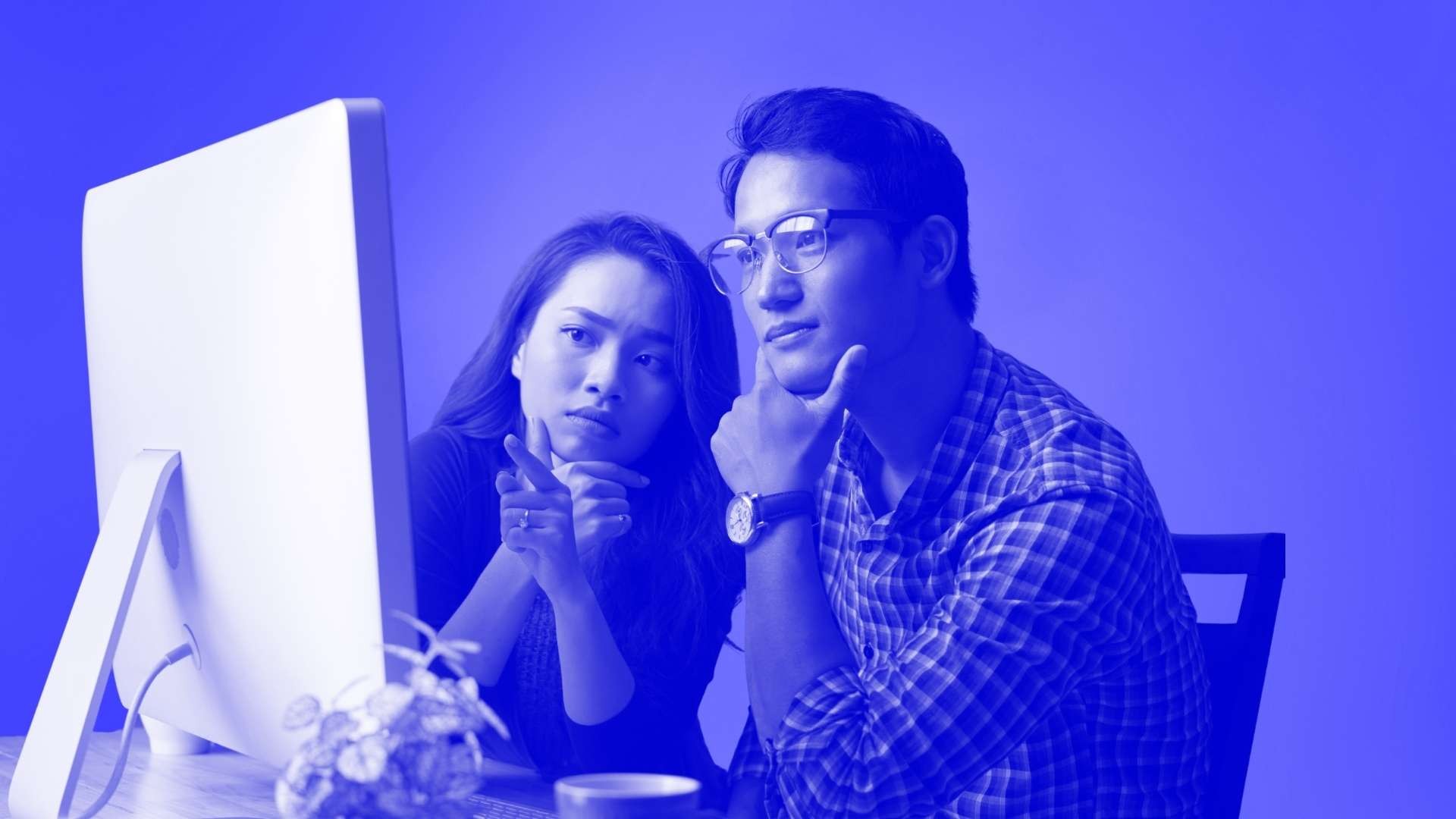 A man wearing glasses and a woman next to him sat in front of a desk looking intently at a computer screen