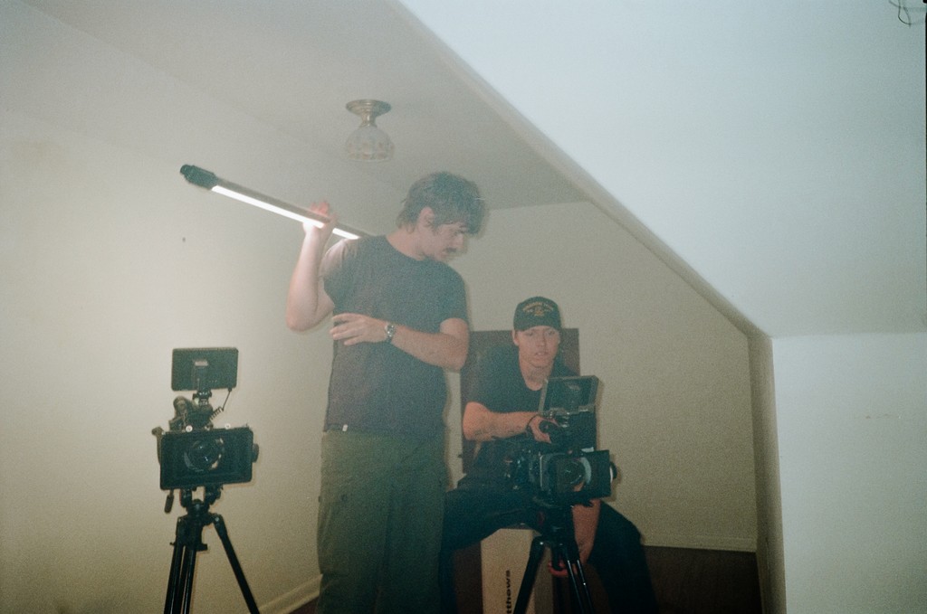 Two men are seen in a relaxed setting, one holding a camera, while a smiling man with a beard sits in the background.