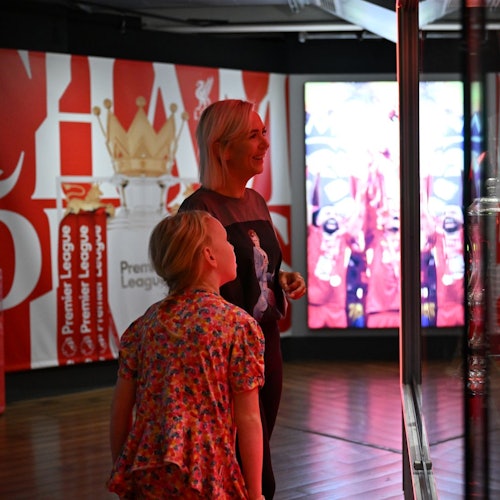 שני אנשים בתצוגת מוזיאון עם פריטי מזכרות מהפרמייר ליג, גרפיקה של כתר גדול ומסכים המציגים תמונות ספורט.