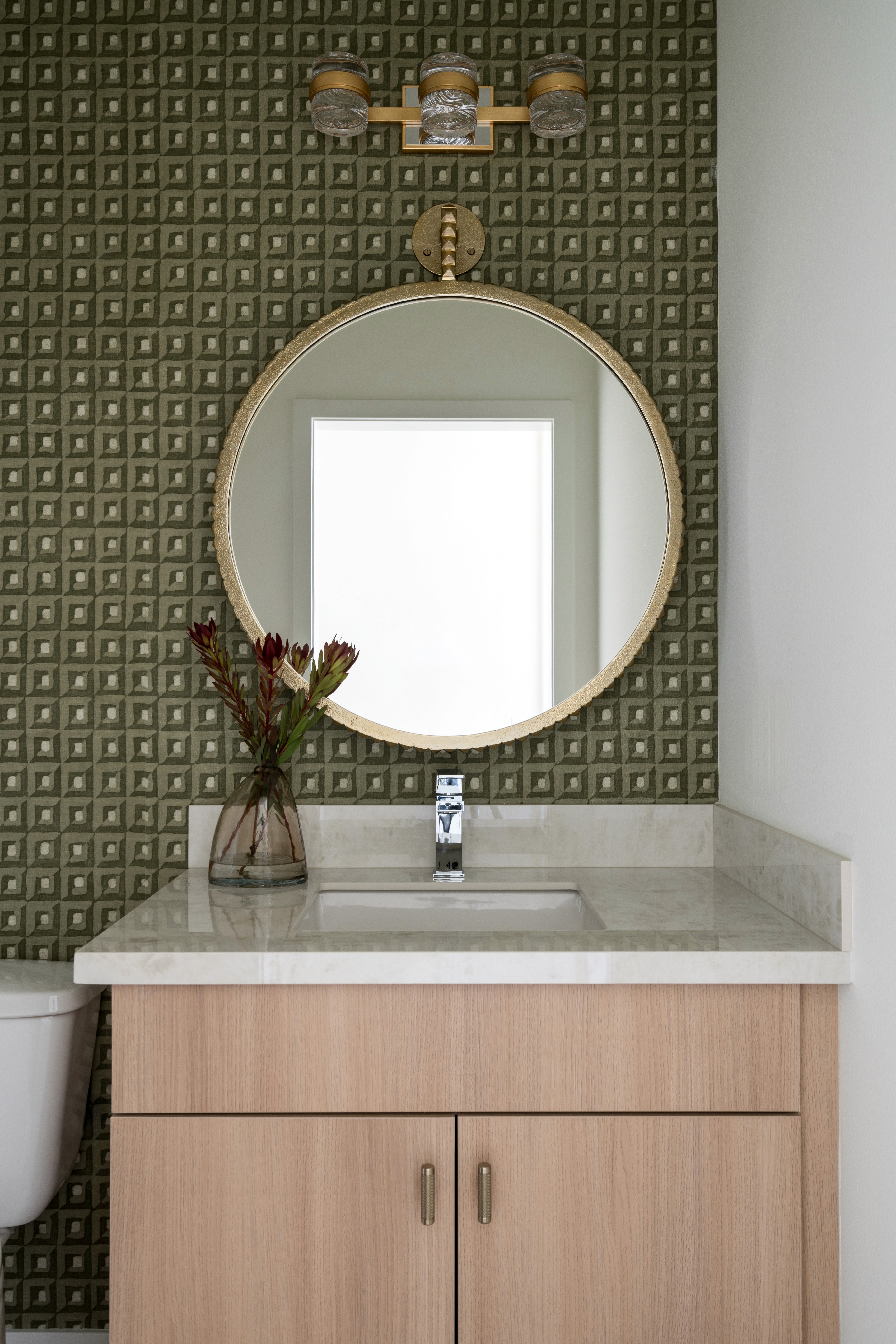 Powder bathroom with circle mirror, green patterned wall paper, clear vase with dried flowers, white countertop, and light wood cabinetry