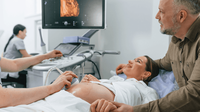 Mulher grávida realizando exame de ultrassom, acompanhada por um homem ao seu lado. Profissional de saúde opera o aparelho de ultrassom, e a imagem do feto aparece na tela.