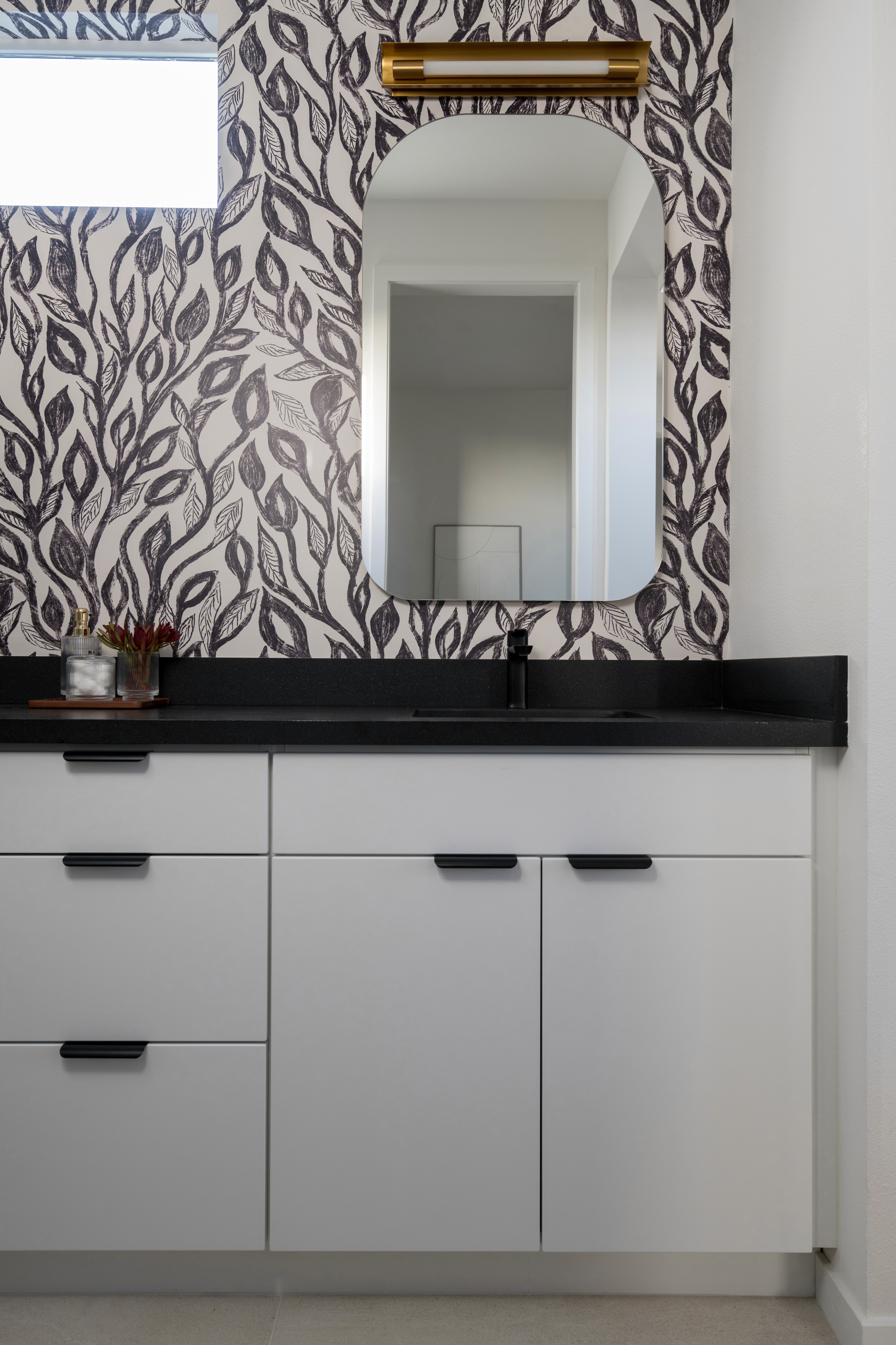 Bathroom vanity with gold framed arched vanity mirror, black and white leaf wall paper, brass sconce lighting, black countertops and flat white cabinets with black hardware