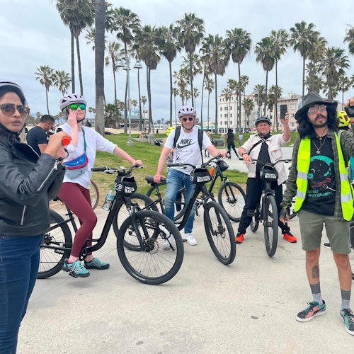 Grupo de ciclistas posando con bicicletas, llevando cascos y ropa informal, cerca de palmeras y un paseo marítimo.