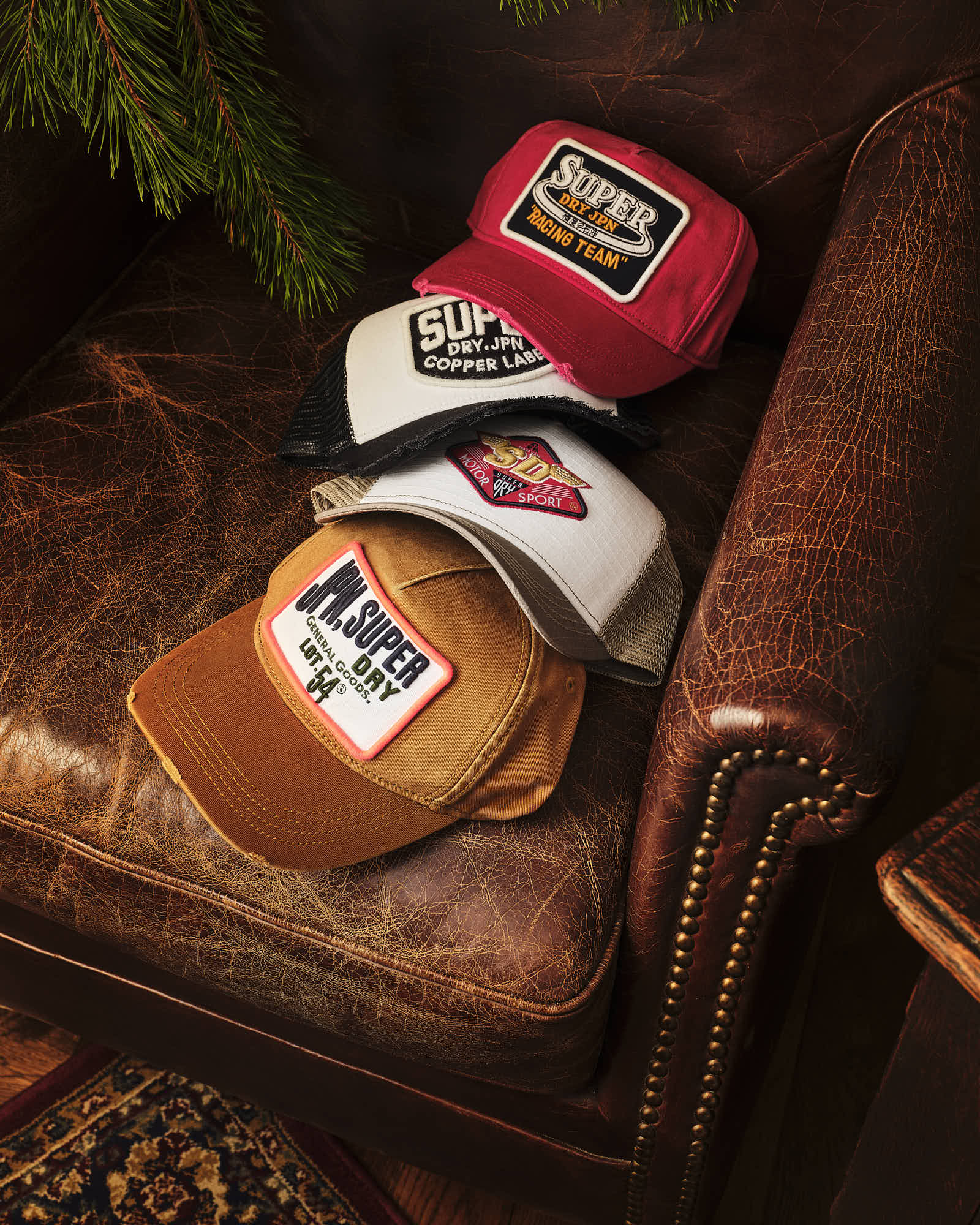 Vintage-style Superdry caps on leather chair with pine branch.