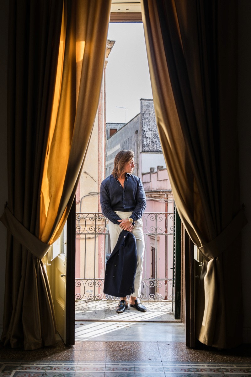 A Don Marrón member stands on an Italian balcony framed by curtains, dressed in a navy suit that reflects refinement and quiet confidence.