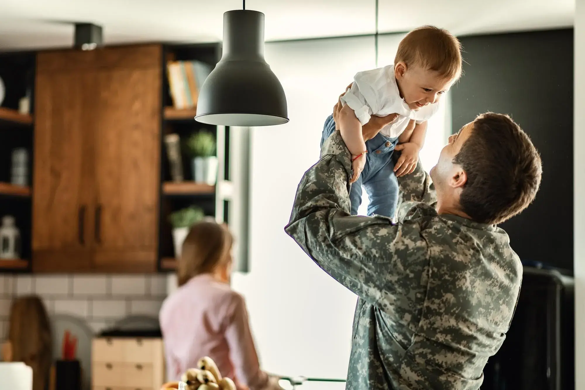 A military parent in uniform lifts their baby joyfully in a bright kitchen, with another parent visible in the background. The image represents family, stability, and the benefits of VA home loans offered by Chris Lewis Home Loans for veterans and military families.