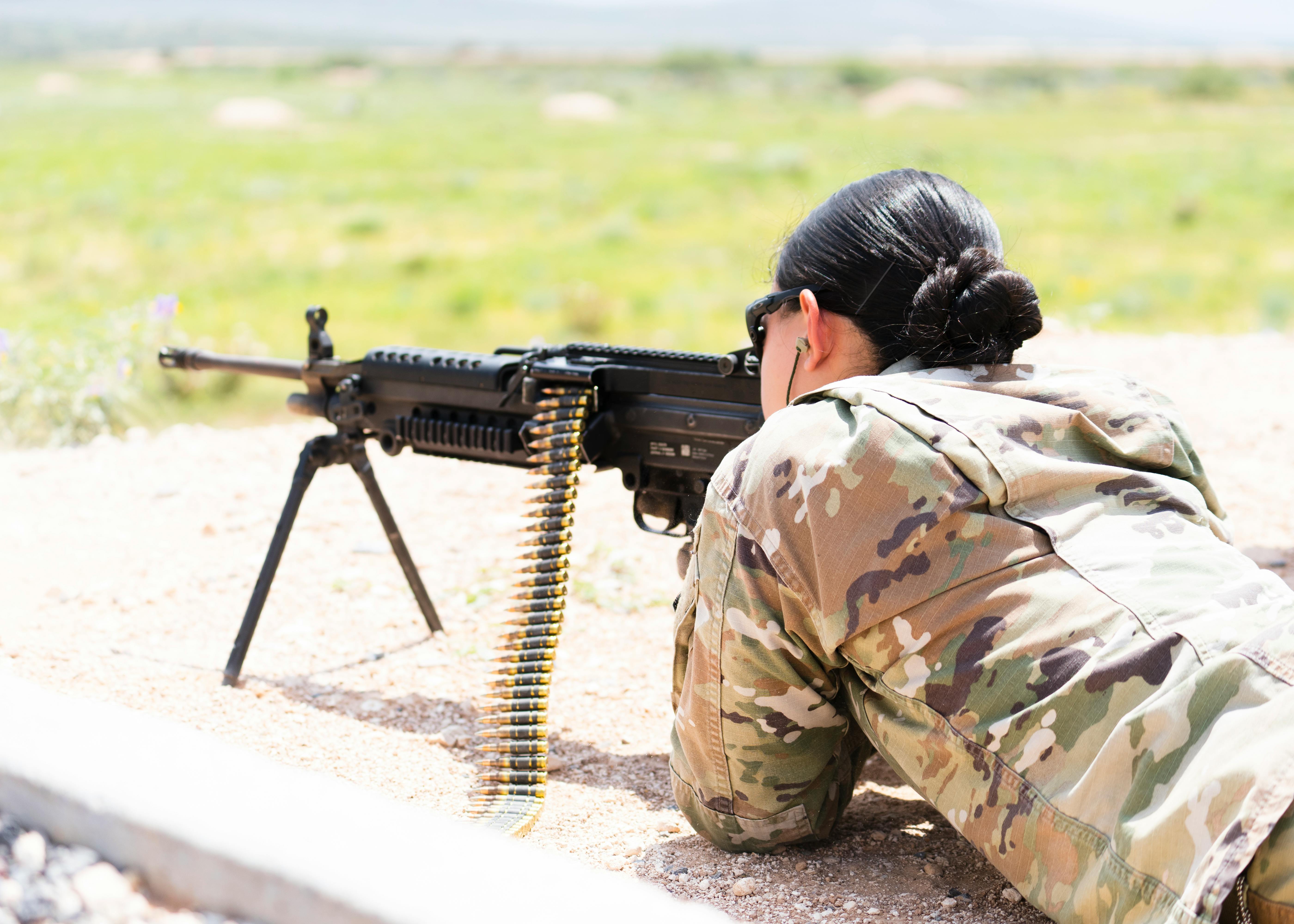 A young woman aims at a target, lying on her stomach, not looking at me, seen from behind. She has black hair styled in a soldier's bun.