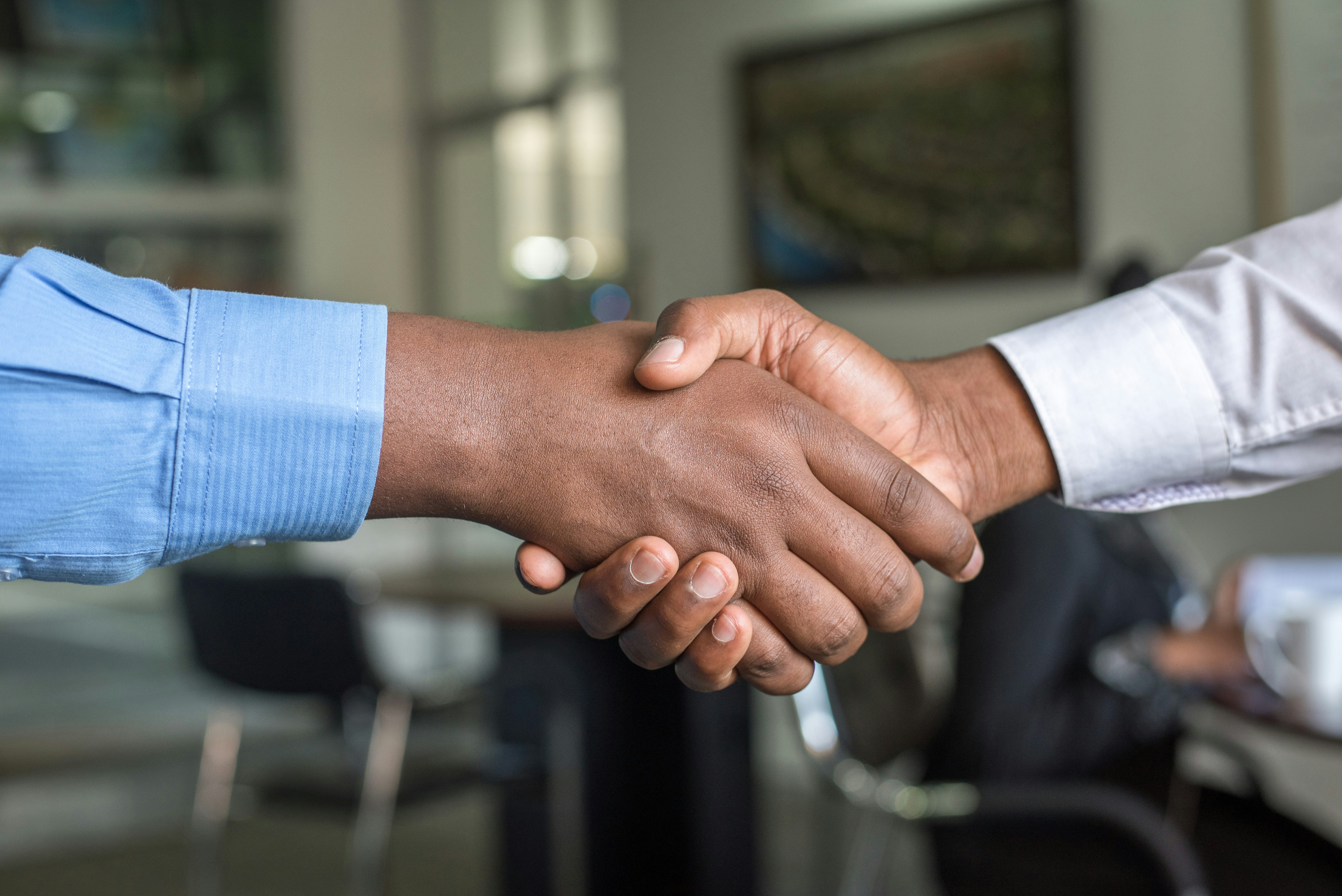 A close-up of two people shaking hands, one wearing a blue shirt and the other in white. The handshake takes place in a blurred office setting, symbolizing partnership, agreement, or collaboration.