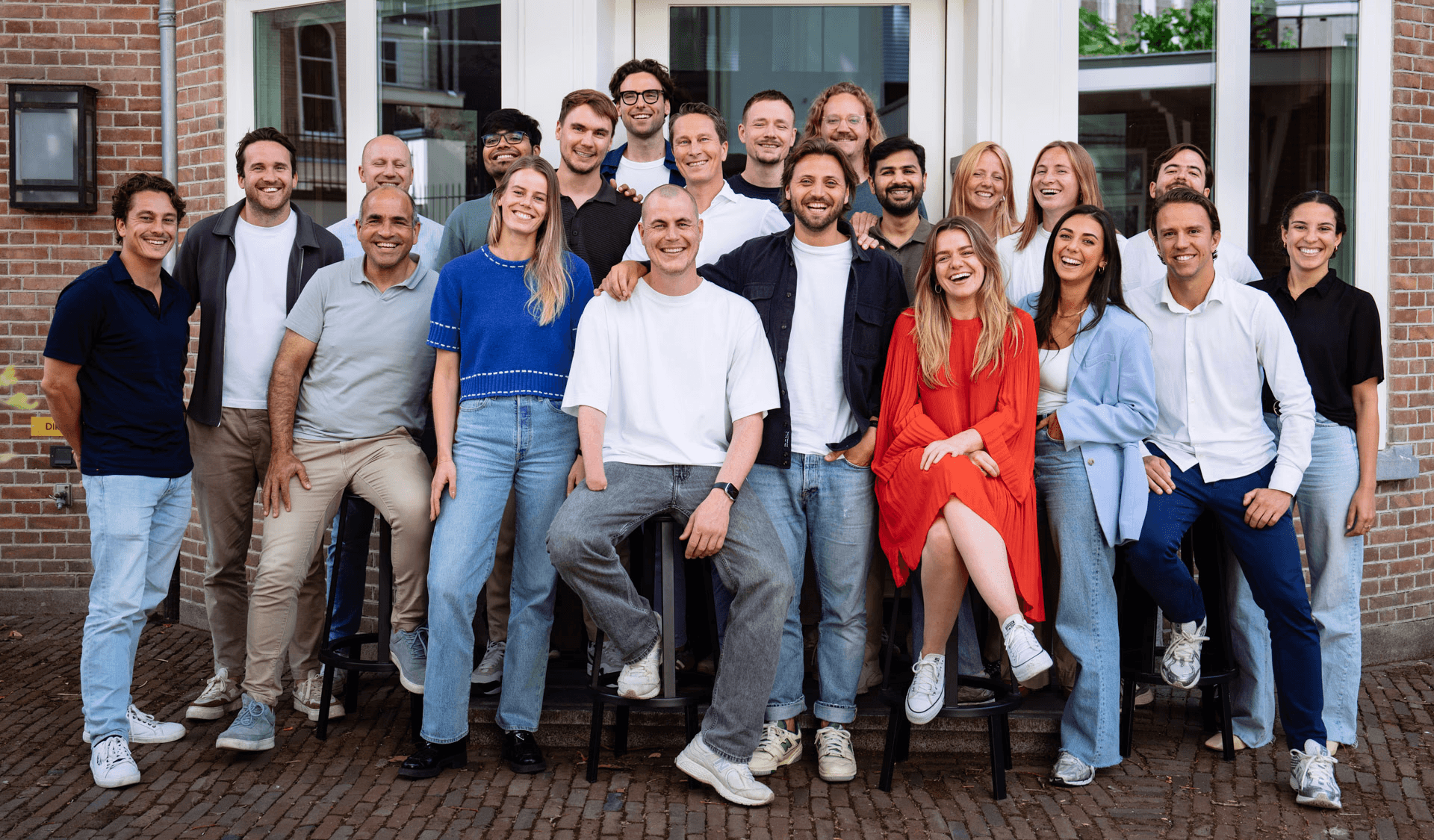 A diverse group of individuals poses together in front of a building, some casually dressed, smiling for the camera.