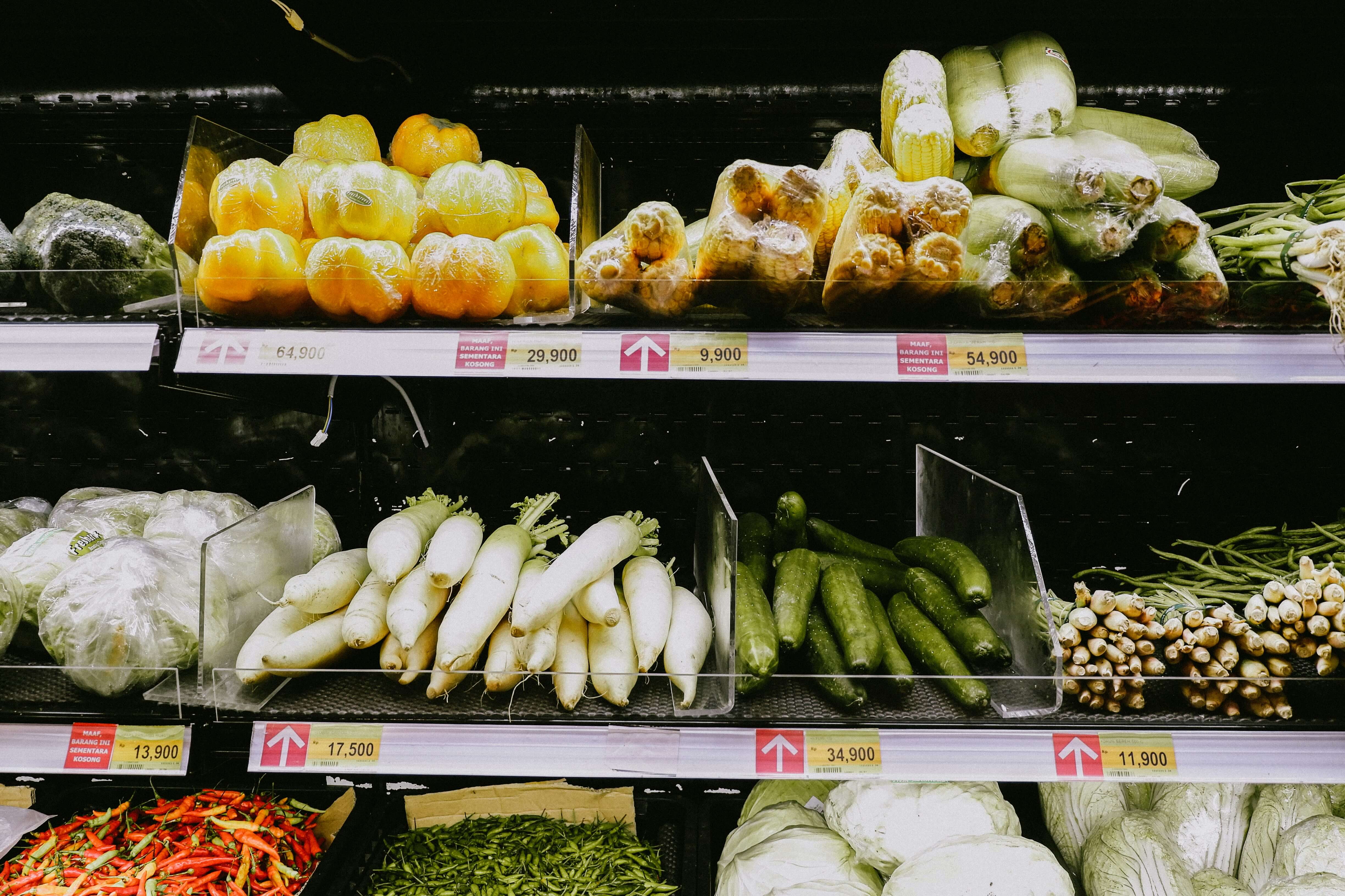 Fresh vegetables displayed on grocery store shelves with visible price labels.