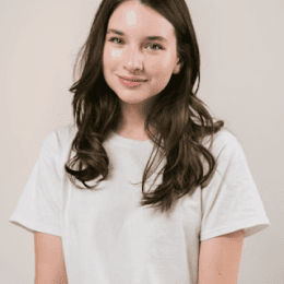 A young woman wearing a white t-shirt, smiling and standing against a neutral background.