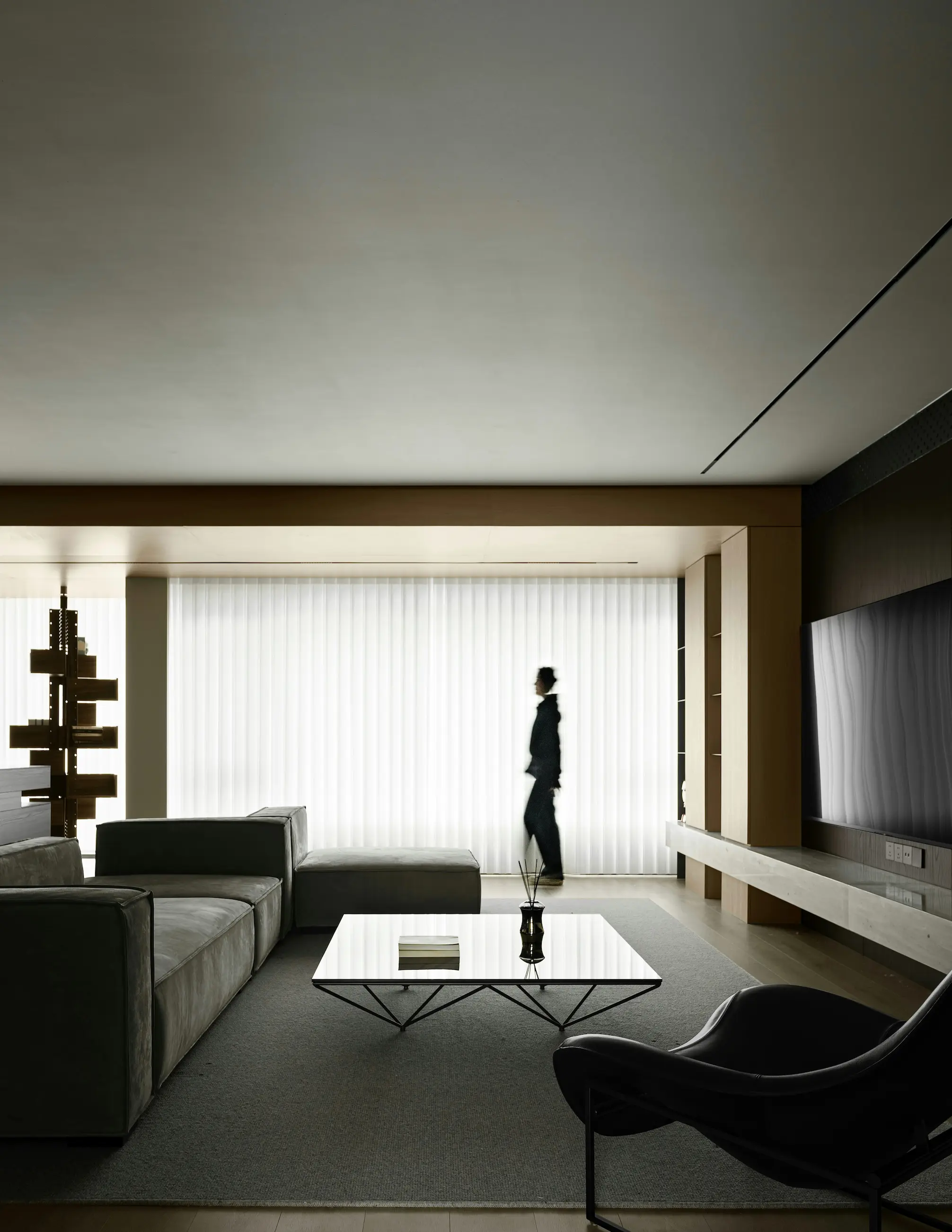 Minimalist living room with a person walking past sheer curtains, large TV and glass coffee table