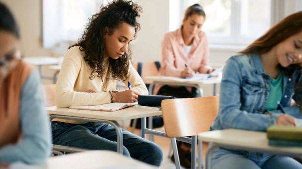 High school students, primarily young women, focused on writing in a classroom during exams or research assignments for RISE Research and STEM summer programs.