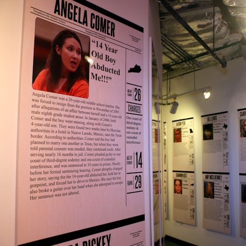 קיר תערוכה עם פוסטרים על מקרים פליליים המציגים תמונות וטקסט על אנג'לה קומר ואחרים בחדר מואר קלות.