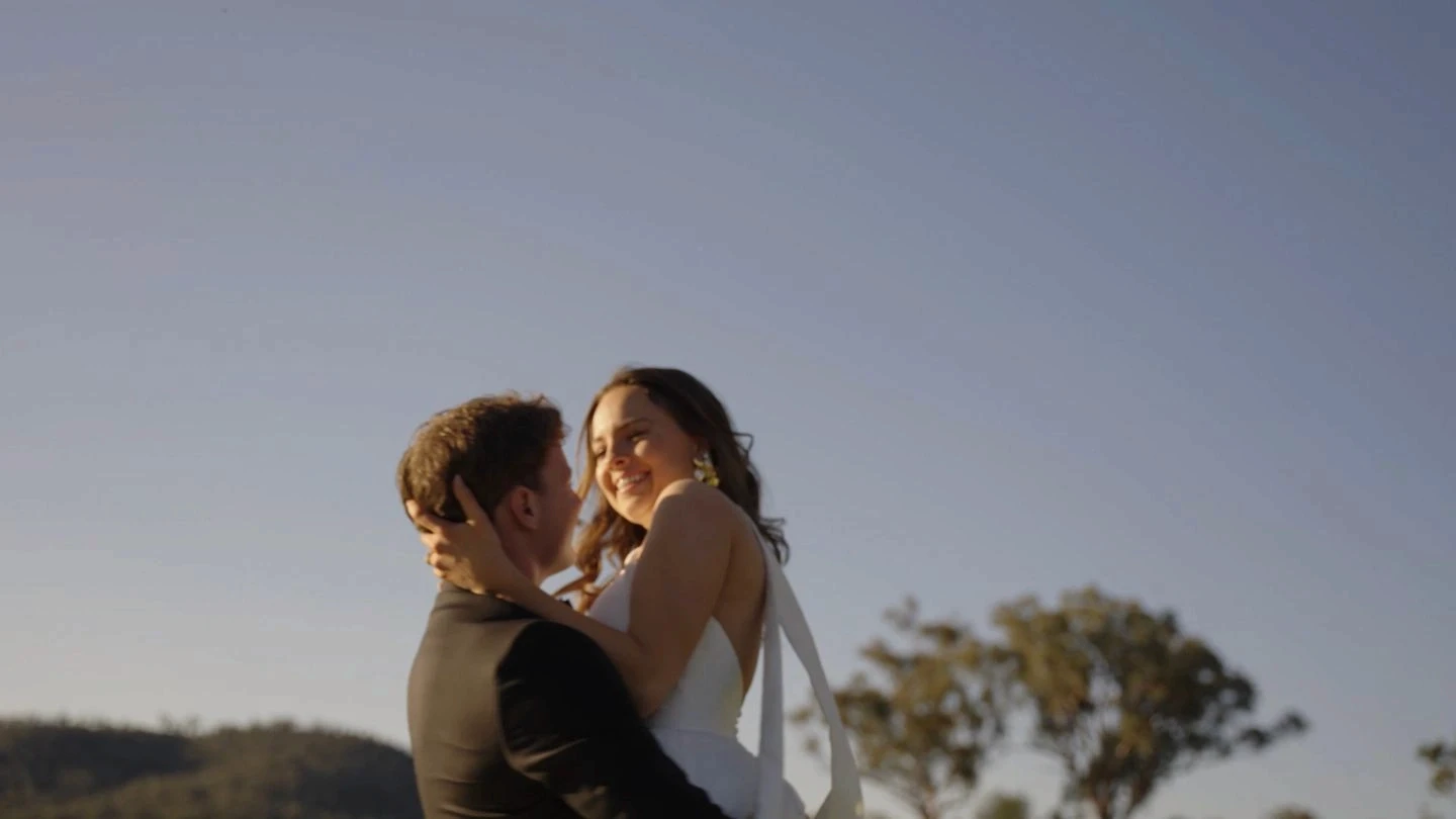 A couple dressed in formal attire share a joyful embrace outdoors under a clear blue sky, with a distant tree and landscape providing a serene backdrop.