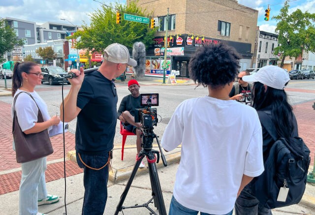 A film crew interviews a man seated on a red chair at a street corner, with cameras and microphones set up on a city sidewalk.