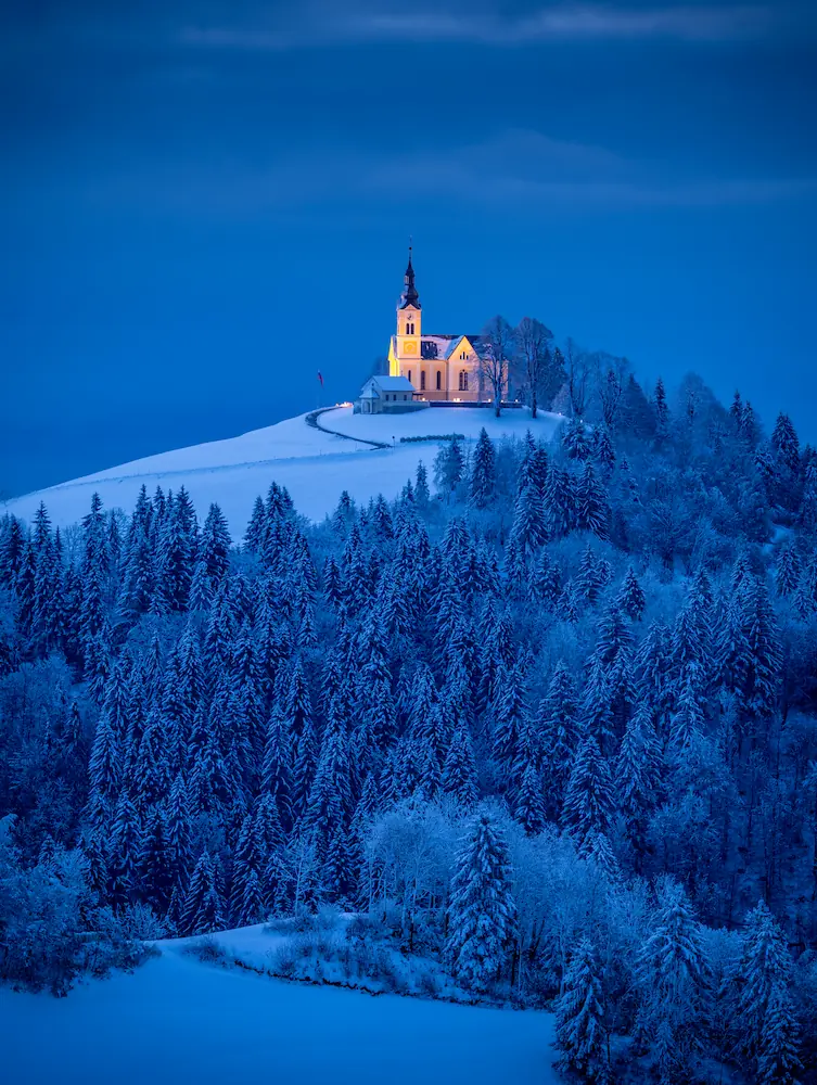 A serene blue landscape features a small church on a hill in Slovenia, surrounded by trees under a twilight sky.