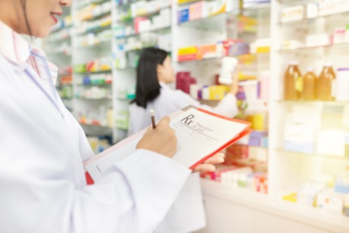 A pharmacist or healthcare worker in a white coat reviewing medication in a pharmacy with medicine shelves in the background