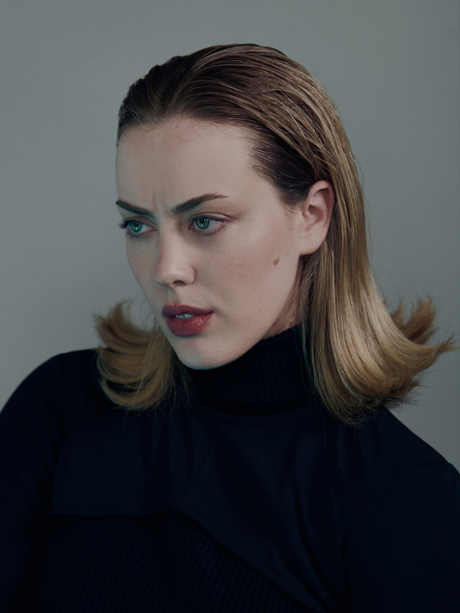An editorial studio portrait of Madison Randolph with green eyes, red lips and blonde hair. The photograph has a moody dramatic tone and expression.