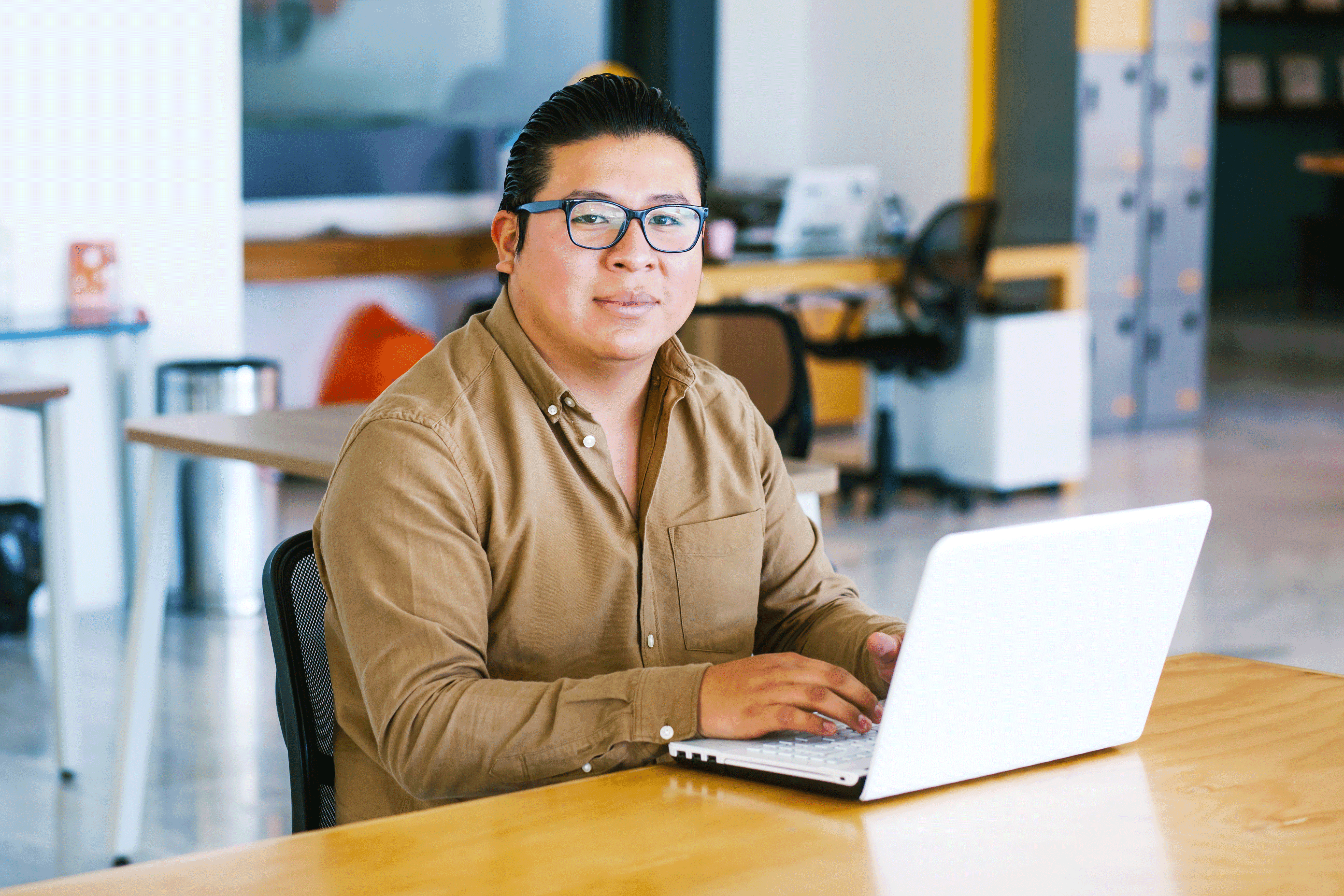A person with glasses and a tan button-down shirt sits at a wooden table, smiling and looking toward the camera while using a white laptop in a modern, open-office setting.