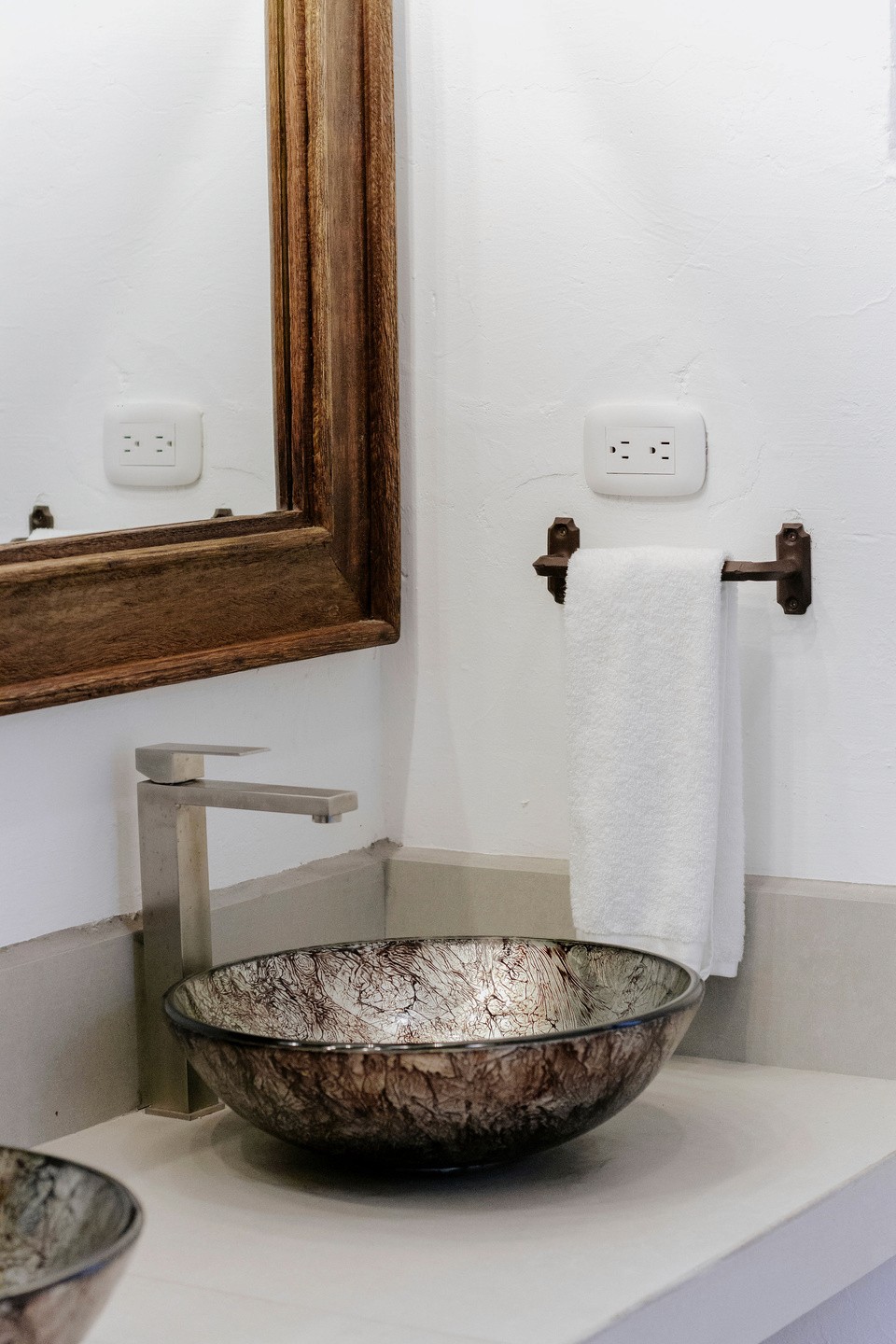A modern bathroom vanity features a stone vessel sink with a sleek, stainless steel faucet, complemented by a rustic wooden-framed mirror and a neatly folded white towel on a metal holder against a white wall.