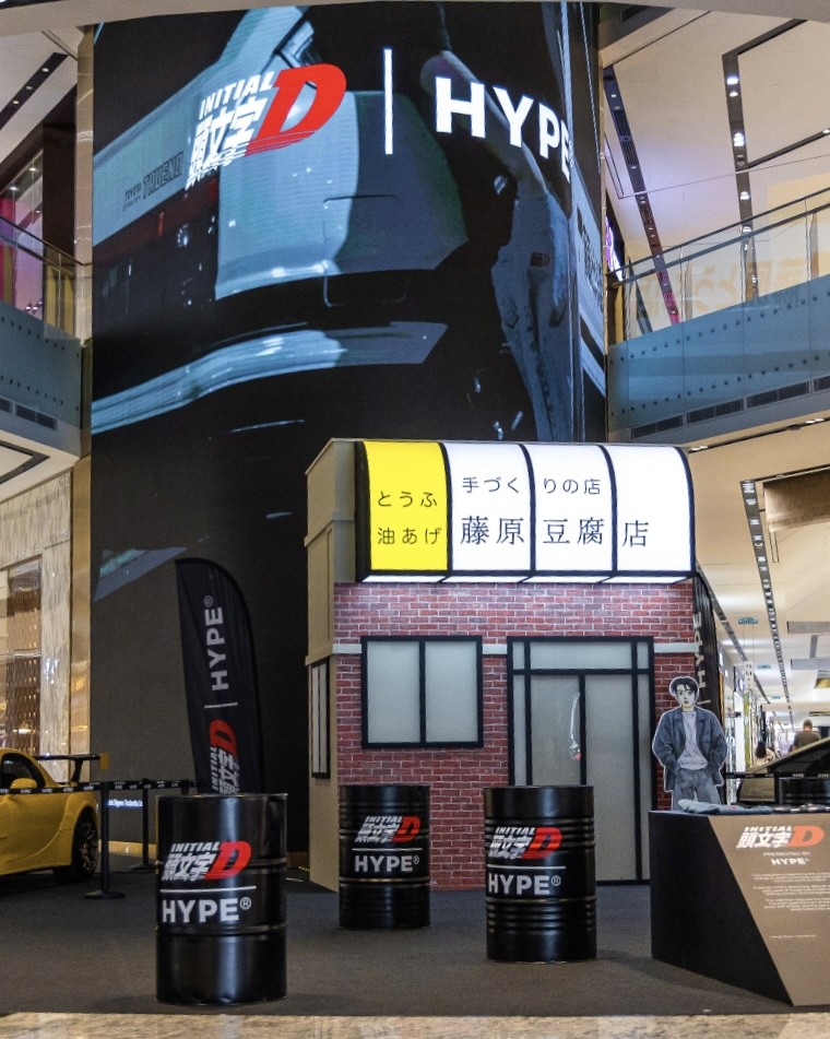 An Initial D x HYPE pop-up display featuring a brick-style structure and promotional materials, set within a mall environment.