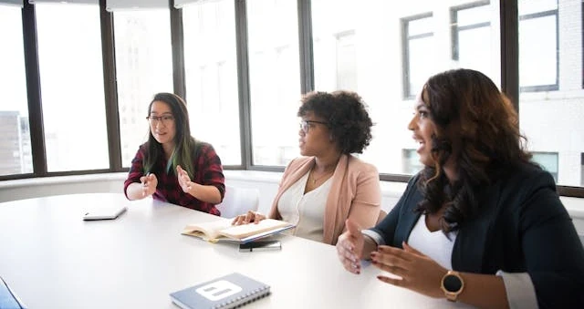 Três mulheres numa sala de reunião conversando à mesa, com cadernos abertos