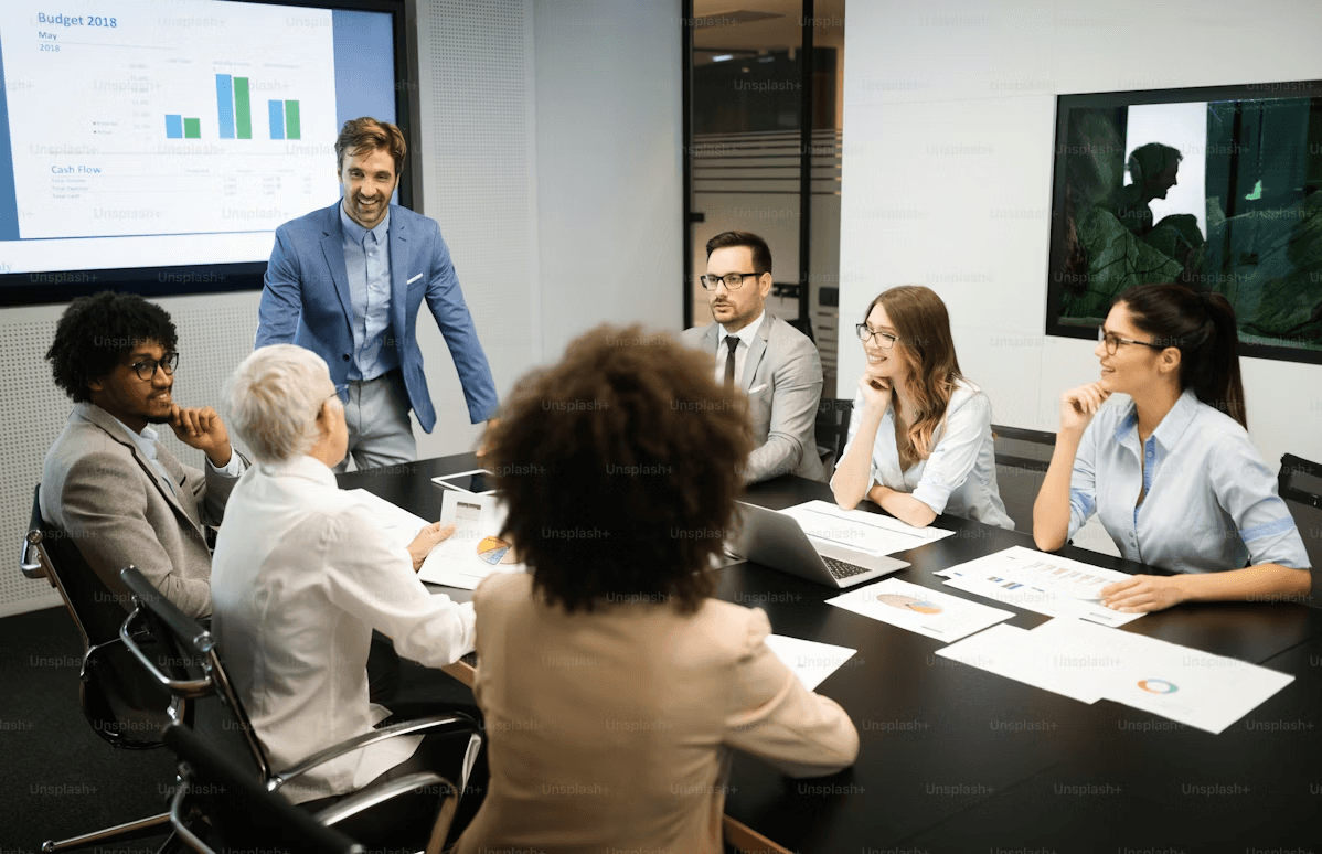 A team meeting in a conference room, with a presenter standing and everyone gathered around a table covered in reports, laptops, and charts.