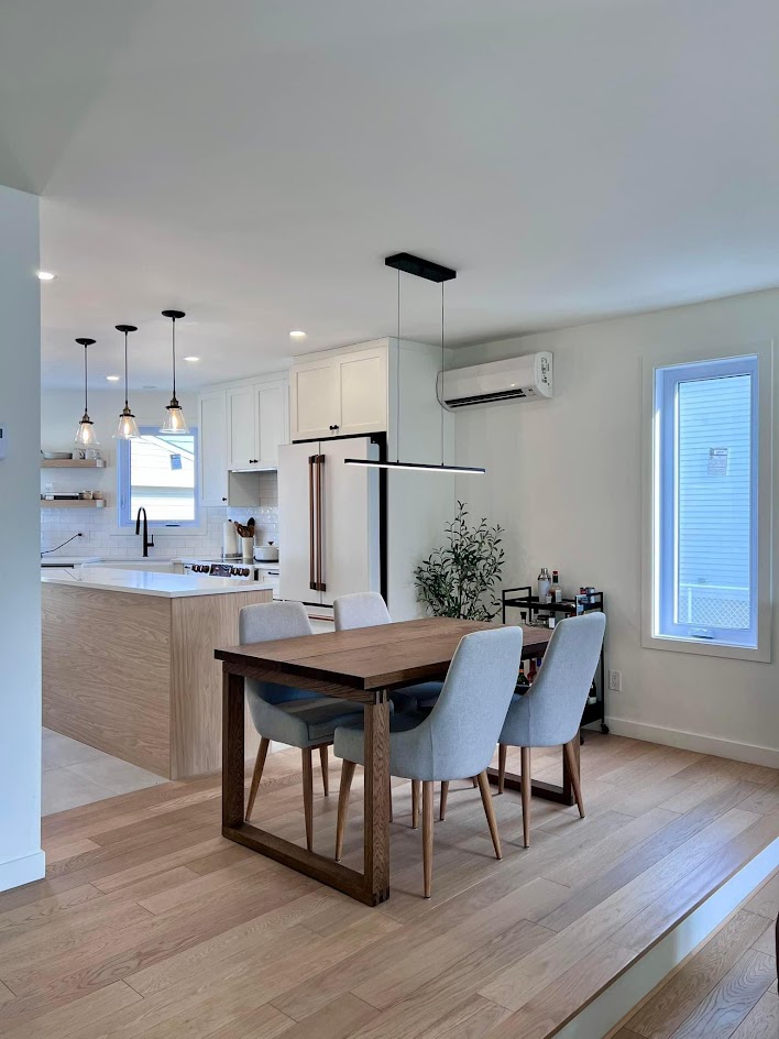  Cuisine blanche et bois blond, équilibrée et lumineuse. Le  contraste entre le blanc et le bois crée une ambiance douce et chaleureuse. L'îlot central, quartz blanc immaculé. Trois suspensions le mettent en valeur. Les électroménagers blancs et leurs accessoires dorés se fondent discrètement dans le décor. Les tablettes en bois près de la fenêtre rappellent le bois de l'îlot. Le dosseret blanc accentue la pureté de la cuisine. Le plancher de bois s'harmonise naturellement avec les teintes de l'espace. La table à manger en bois plus foncé ajoute un beau contraste. Un luminaire linéaire minimaliste complète l'ensemble. Des chaises en tissu apportent une touche de douceur.