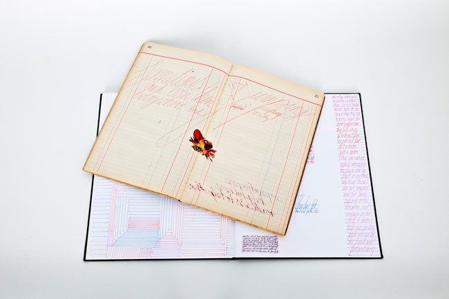 An open book lies on a white surface, displaying handwritten text in red ink on its cream-colored pages. A small, embroidered red flower is positioned near the center. A black spine runs along the right edge of the book, and a pink-and-white geometric pattern is visible on the lower left page. The pages show signs of age, with slight discoloration and wear.