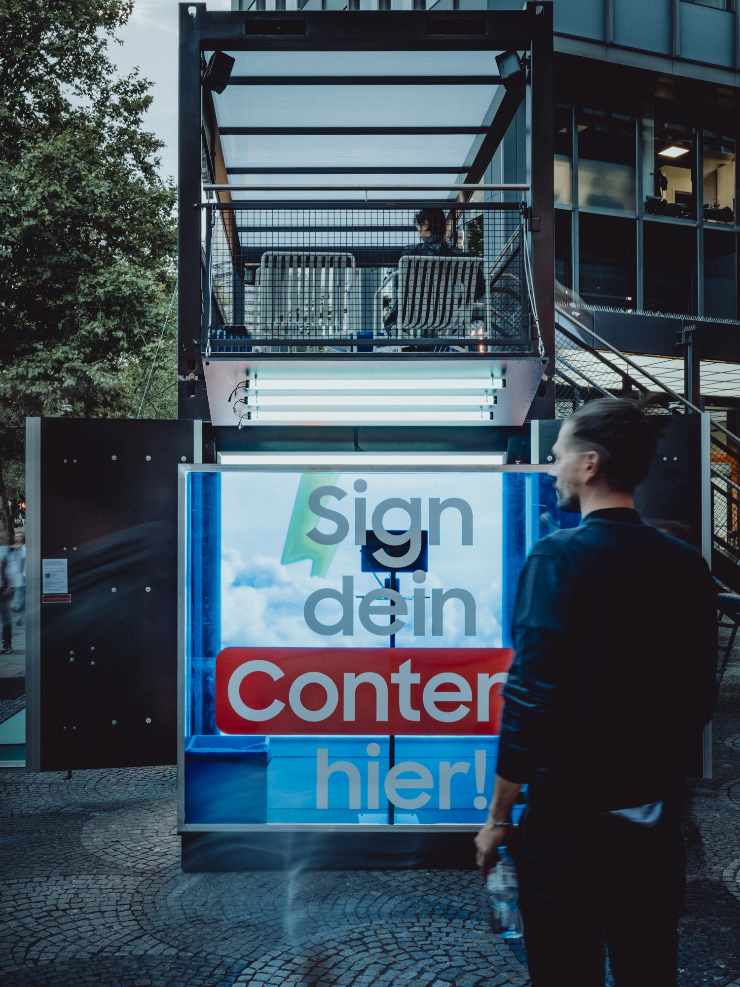 Illuminated facade panel of the SAMSUNG x TIKTOK module with Sign dein Content hier lettering and visitors on the roof terrace above.