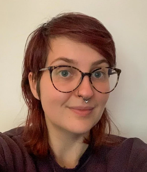 A portrait‑style photograph of a person shown from the shoulders up against a plain, light‑coloured background. They have dark red, shoulder‑length hair worn loose and are wearing glasses with dark frames and a dark, long‑sleeved top. The image is softly lit and closely cropped, giving the feel of a casual, indoor head‑and‑shoulders photo.