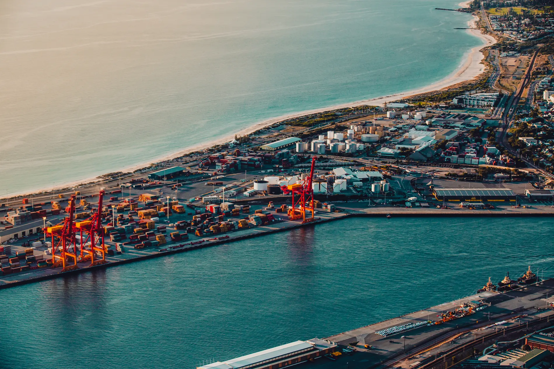 Aerial at dusk of a container port and its red cranes, with a long beach curving away to a coastal town