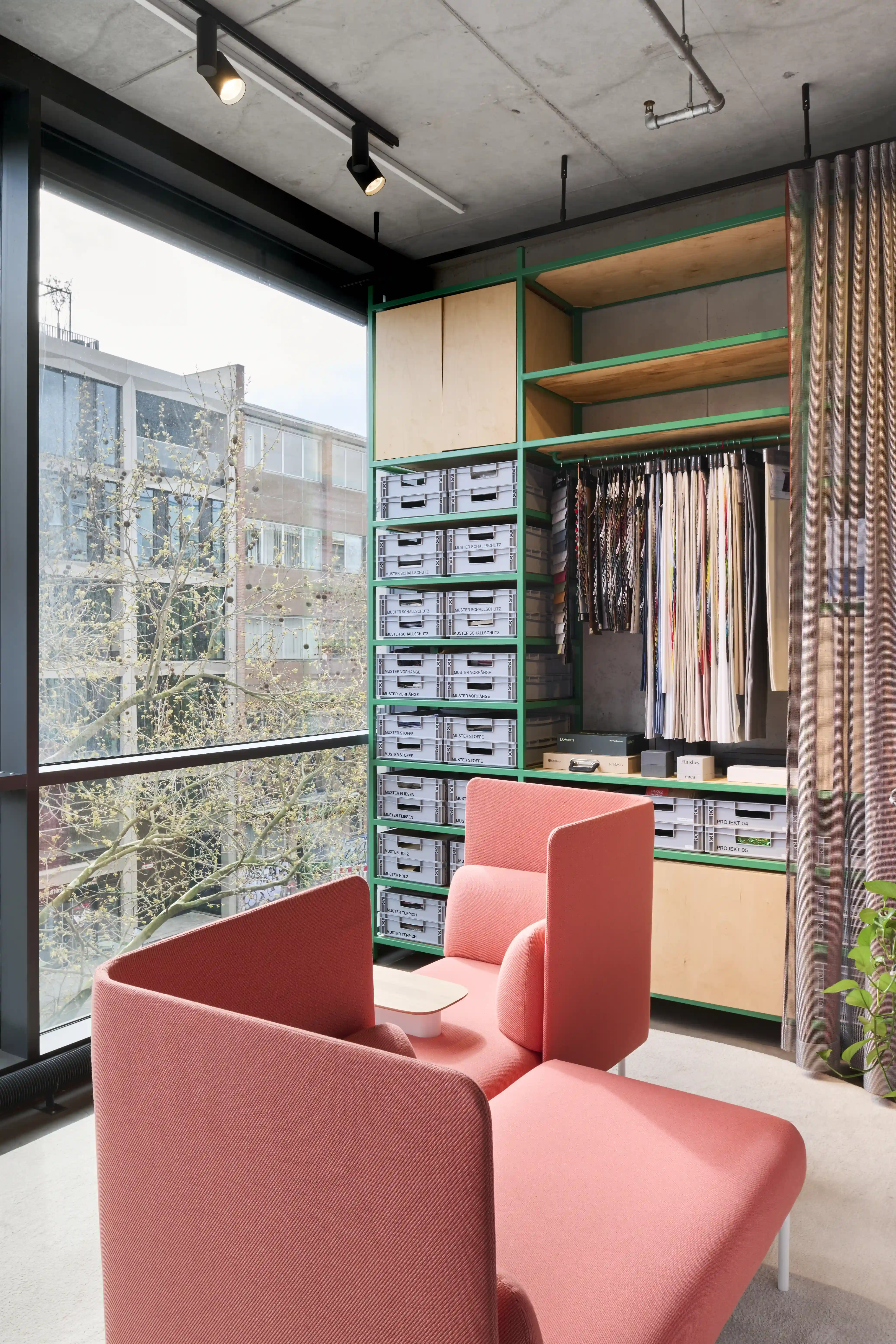 Material archive in the UNDPLUS SHOWROOM with a green steel shelf holding grey sample crates and hanging fabric swatches, in front of two coral armchairs by the window.
