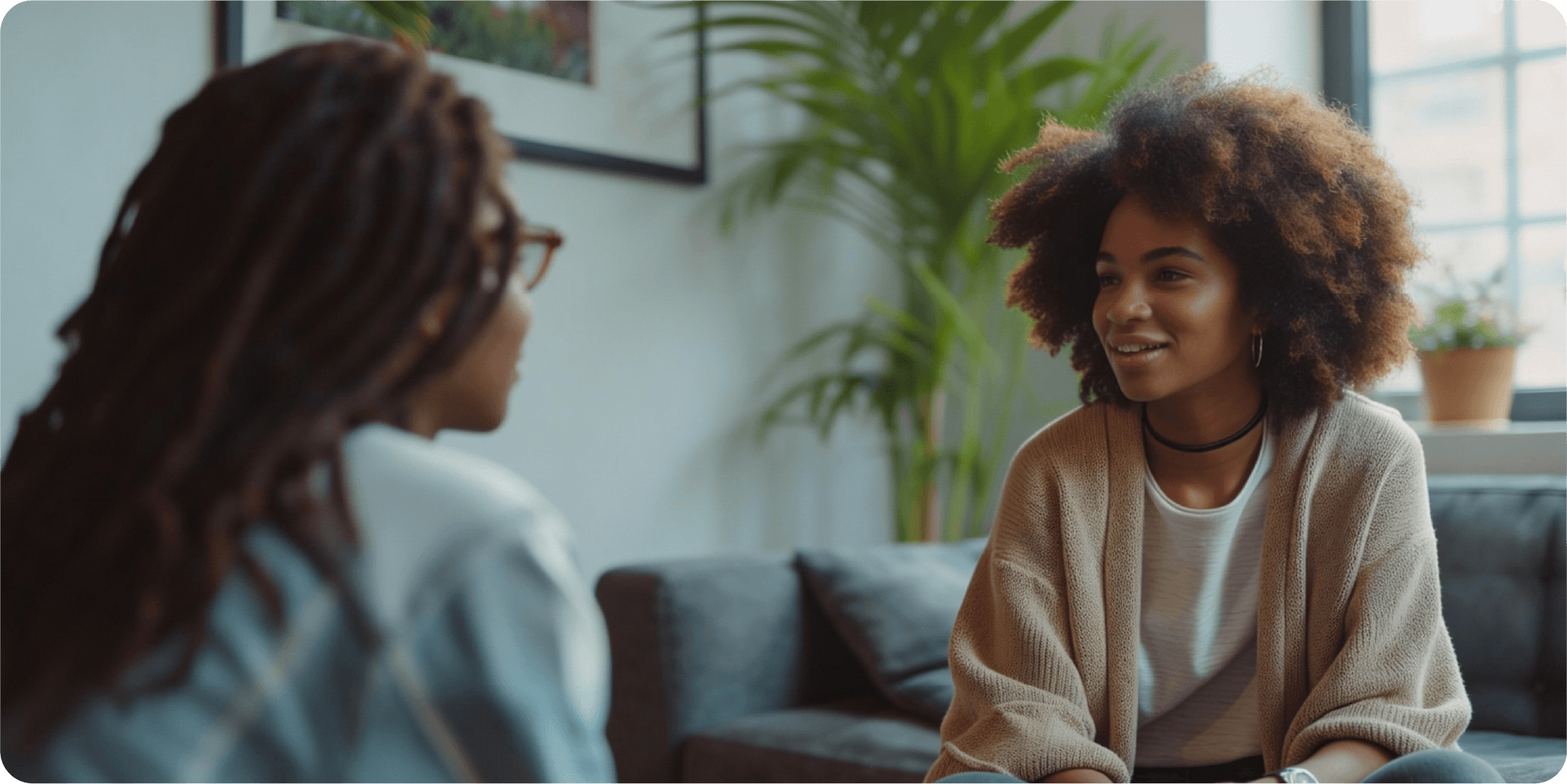 Two young women are sitting on a couch, a therapist and her patient. They facing each other, making eye contact and engaged in conversation. The woman on the right is the patient and she has dark, curly hair and is wearing a beige cardigan over a white shirt and a black choker necklace. She is smiling and looking at the therapist woman. The woman on the left is the therapist and she has dark dreadlocks and is wearing glasses and a light blue jacket. Her back is mostly to the camera, and her face is not visible. The background is a living room with a large window, plants, and a framed picture on the wall. The lighting is soft and natural, creating a warm and inviting atmosphere.