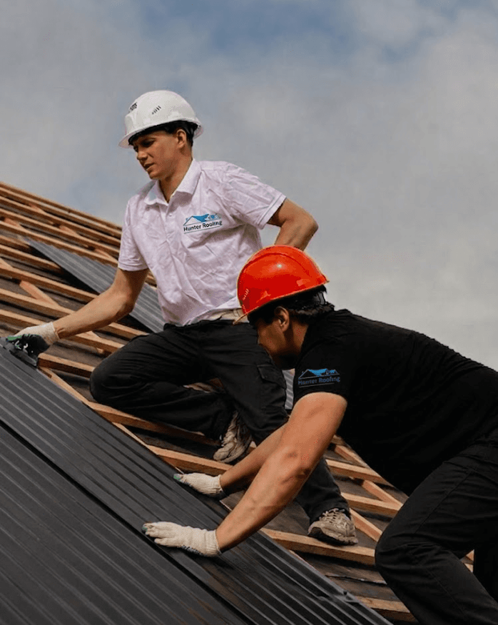 Two construction workers wearing hard hats install dark roofing sheets on a wooden roof under a cloudy sky.