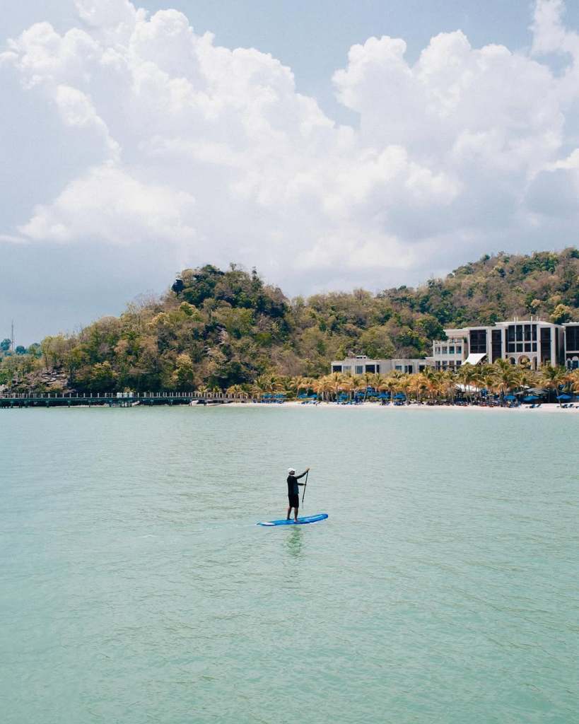 A person paddleboarding in calm waters near The St. Regis, Langkawi, with a backdrop of lush greenery and hotel buildings.