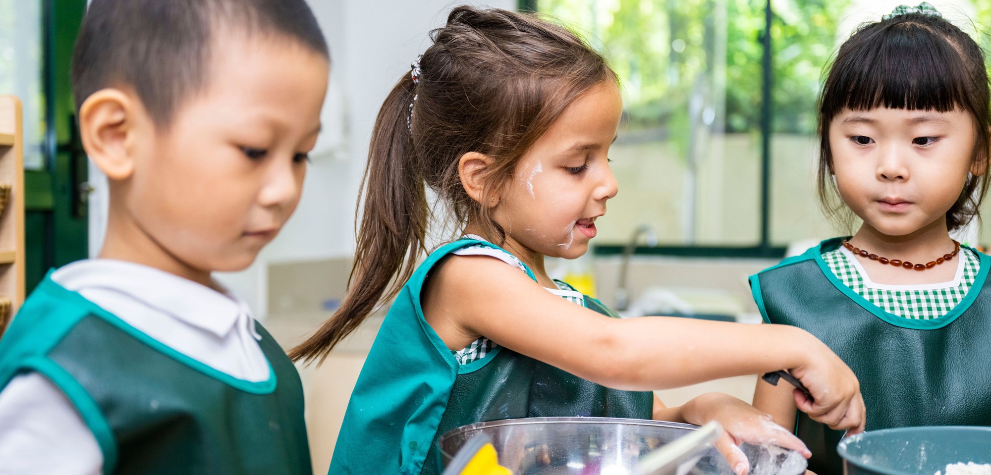 Three early years children wearing green aprons whilst baking