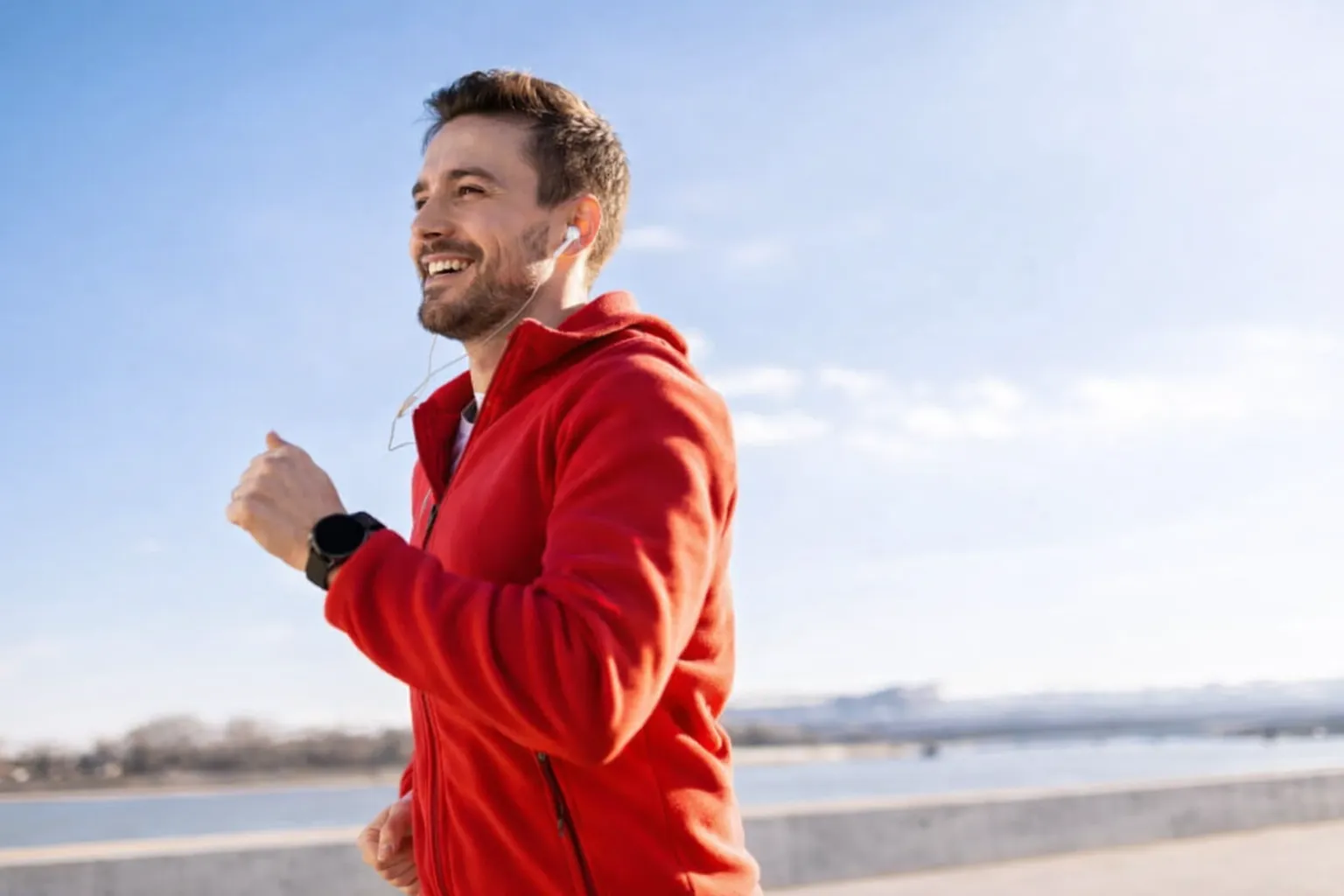 Man in a red jacket jogging outdoors by the water on a sunny day, wearing earbuds and a smartwatch