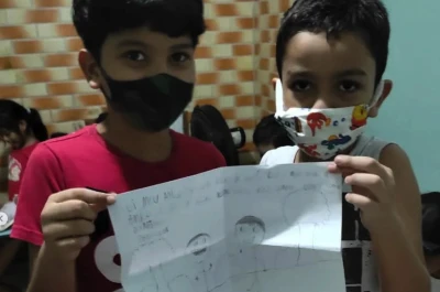 Dois meninos usando máscaras mostram seus desenhos em folhas de papel, orgulhosos de suas criações.