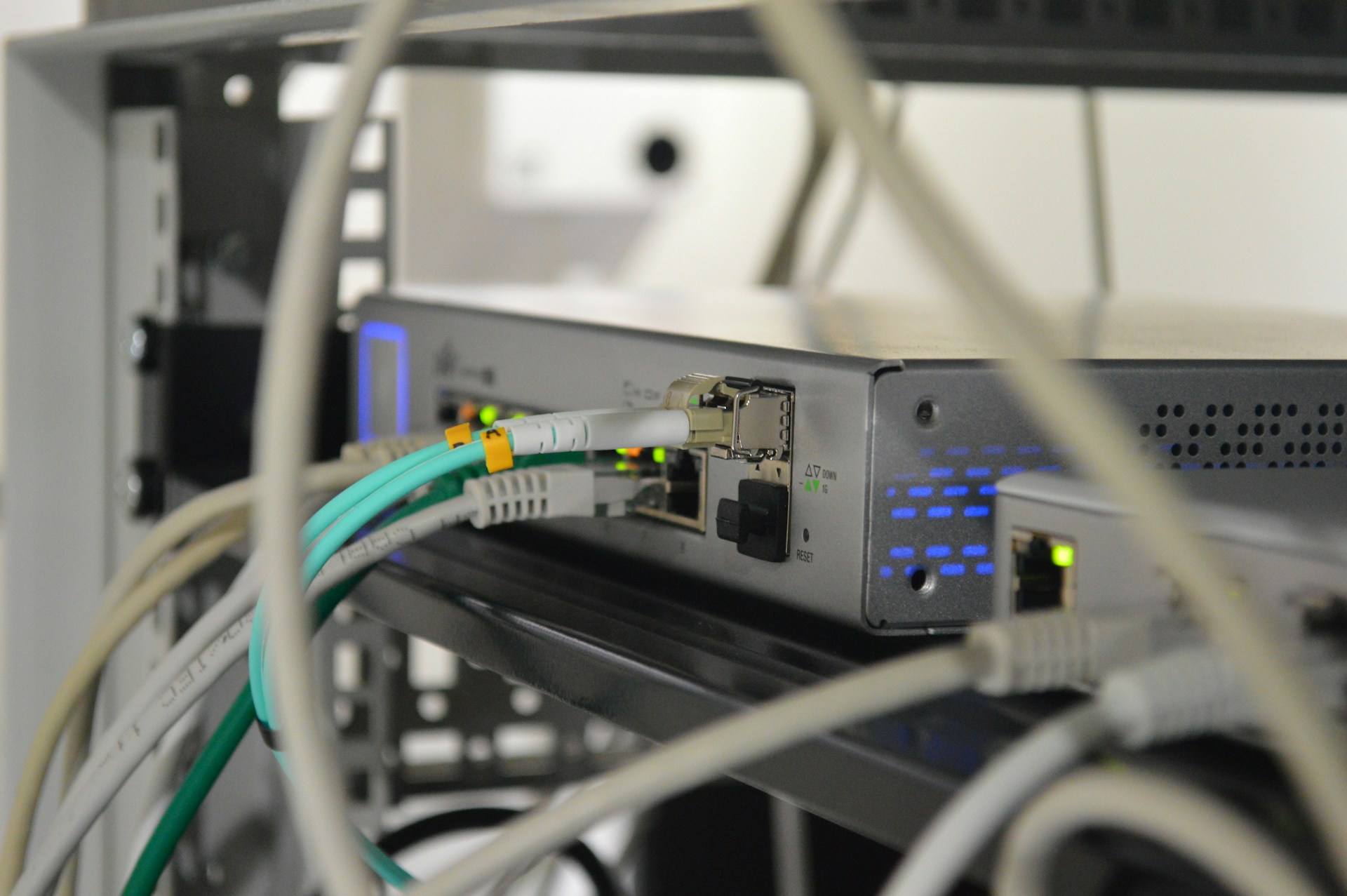A close-up shot of a rack-mounted network switch or router with multiple Ethernet cables (white, teal, gray) plugged into the front ports, indicated by glowing green and blue status lights.