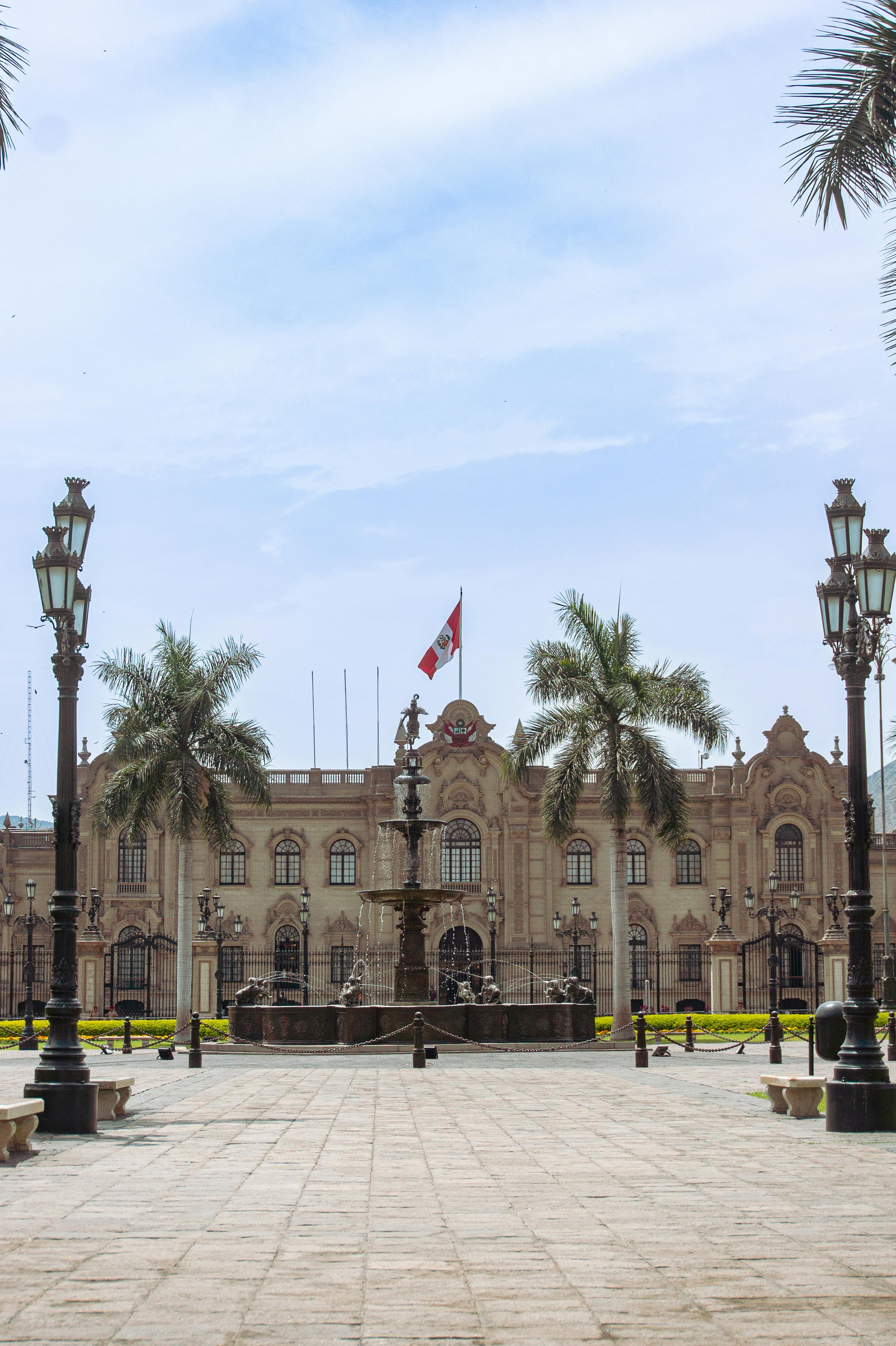 The Government Palace in Lima, seen from the Plaza de Armas. A large ornate fountain and palm trees are in the foreground, with the Peruvian flag flying atop the grand colonial-style building.