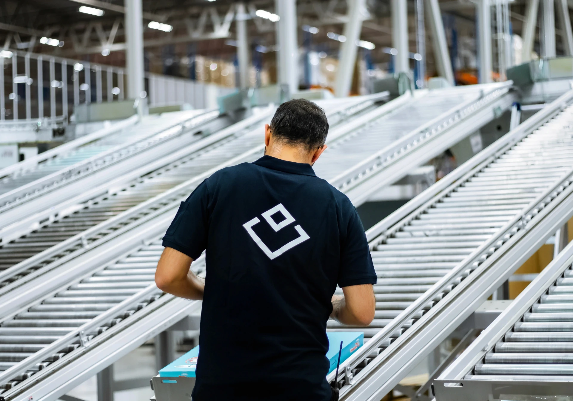 En man i svart skjorta med logotyp arbetar vid transportband på en fabrik, nära bild.